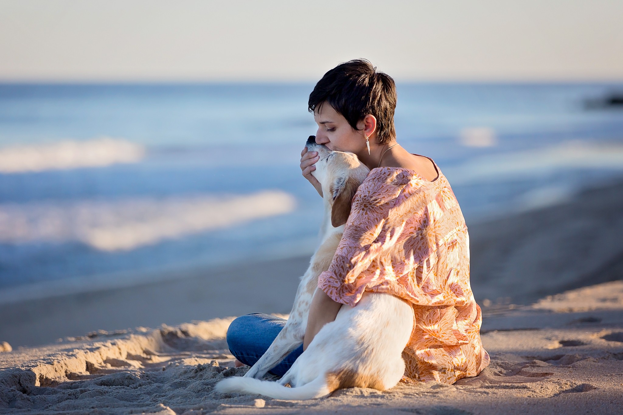 Child sitting on the beach holding a dog, with ocean waves in the background.