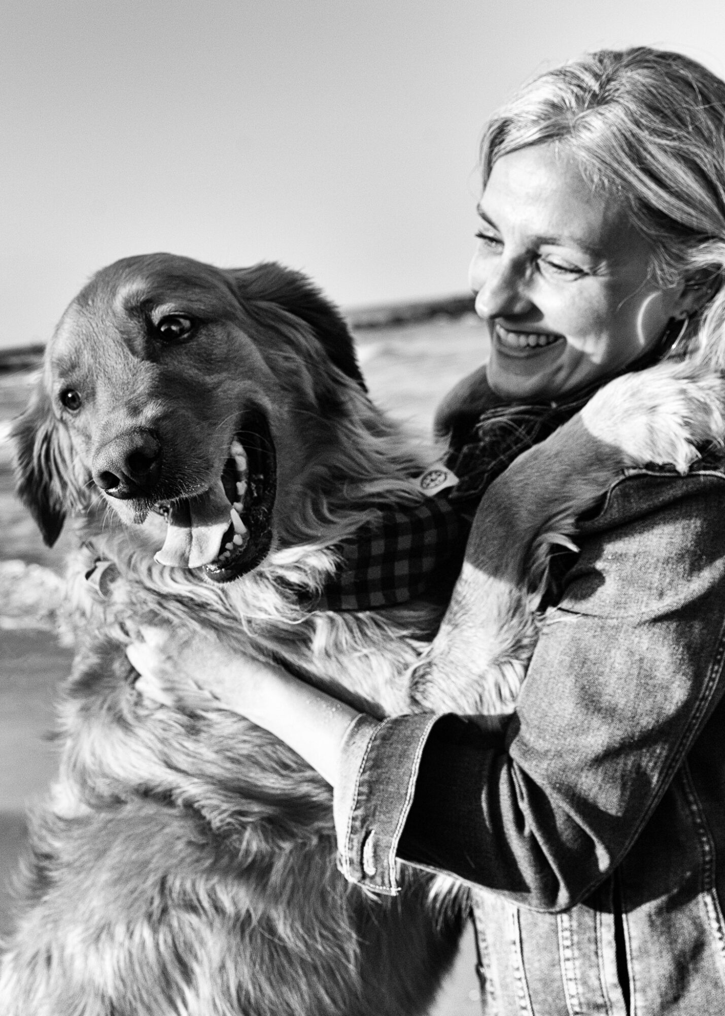 Woman smiling and hugging a dog outdoors, both facing each other, in black and white.