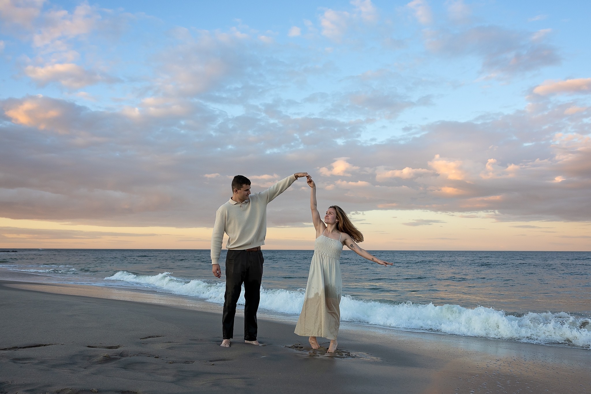 A man and woman holding hands and dancing on the beach near the water, with a cloudy sky and ocean in the background.