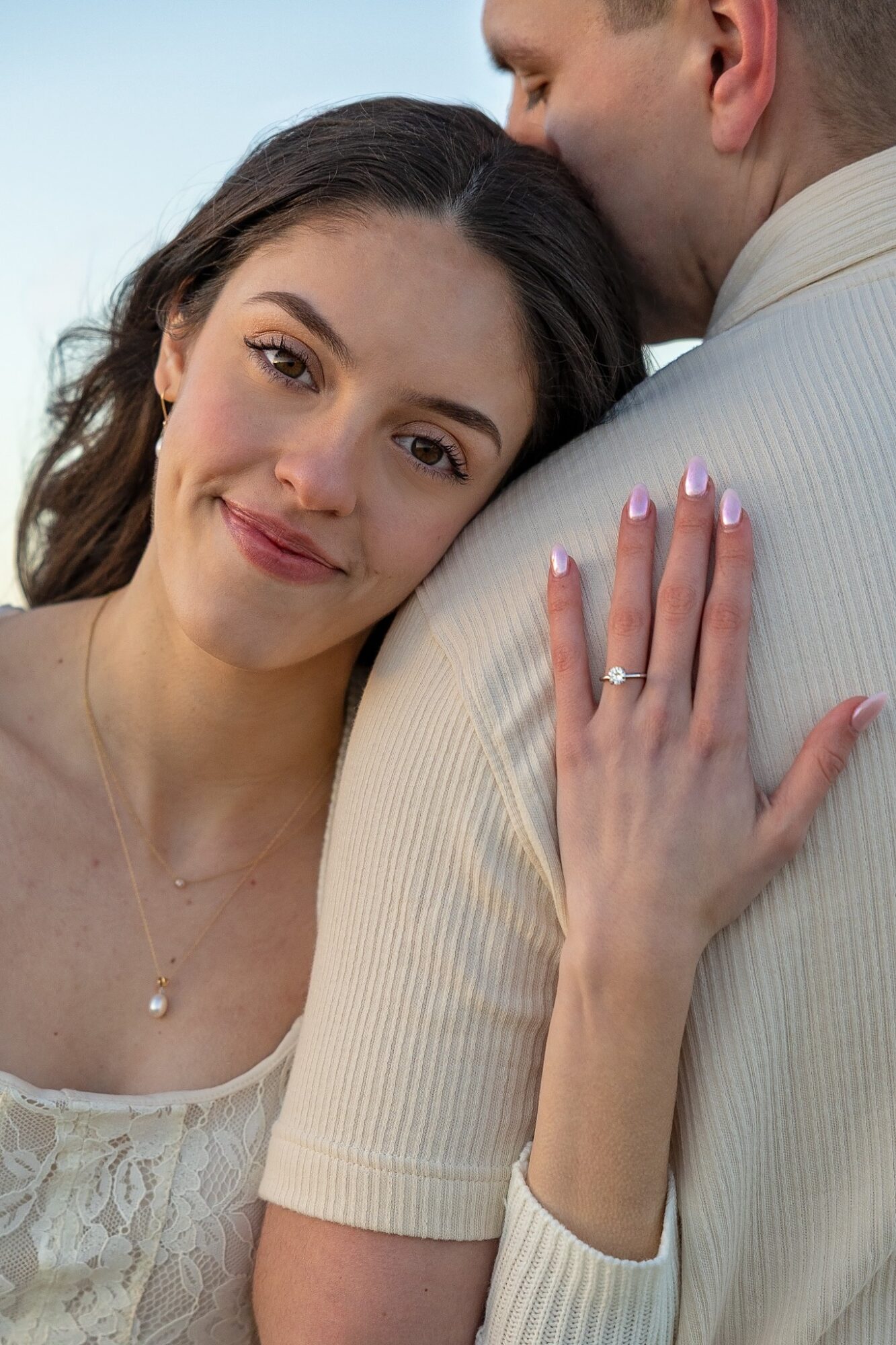 Woman with dark hair and a ring on her finger leans on a man's shoulder, smiling softly, with her hand on his shoulder.