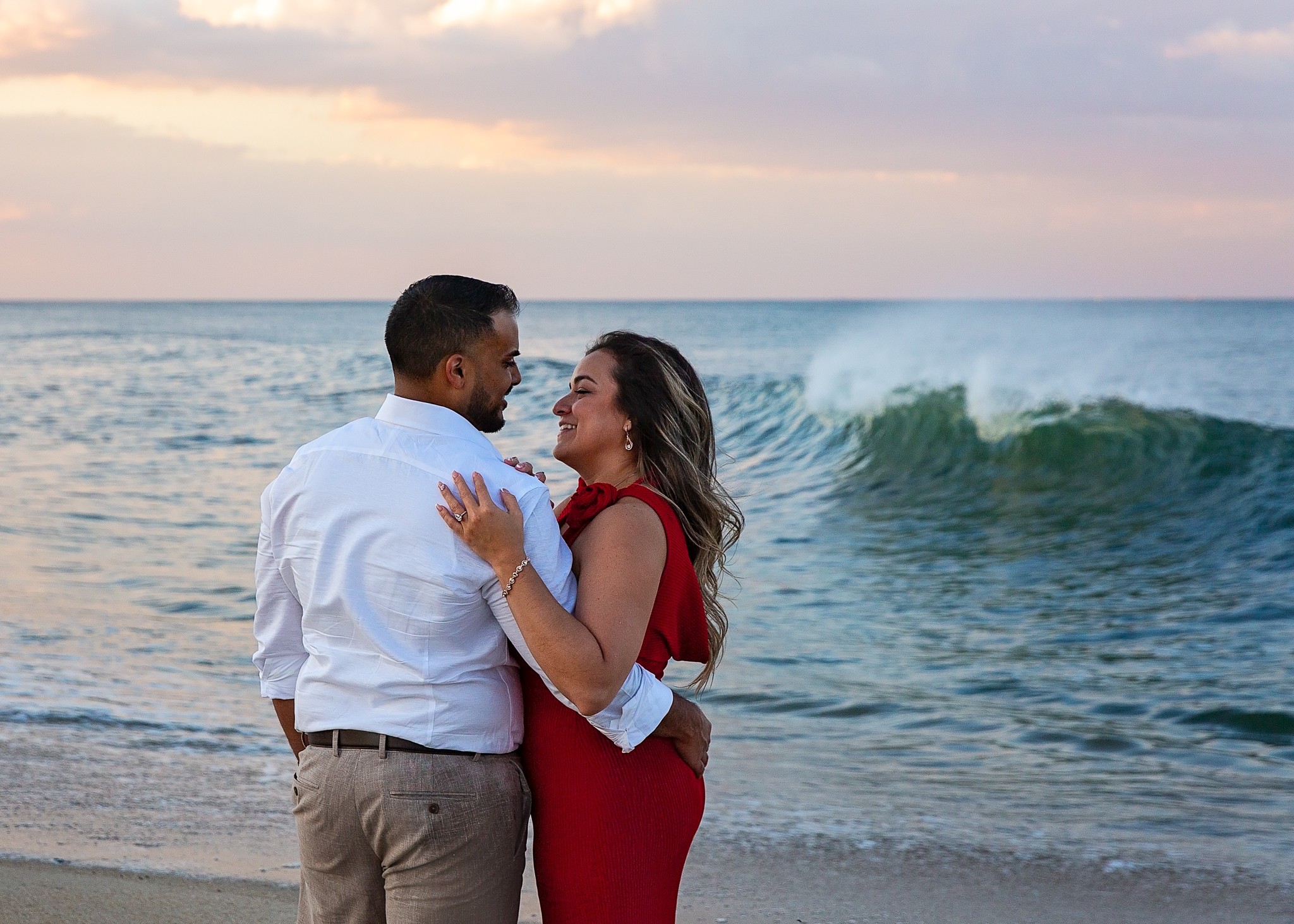 A couple stands on the beach near the ocean, embracing with waves and a sunset sky in the background.