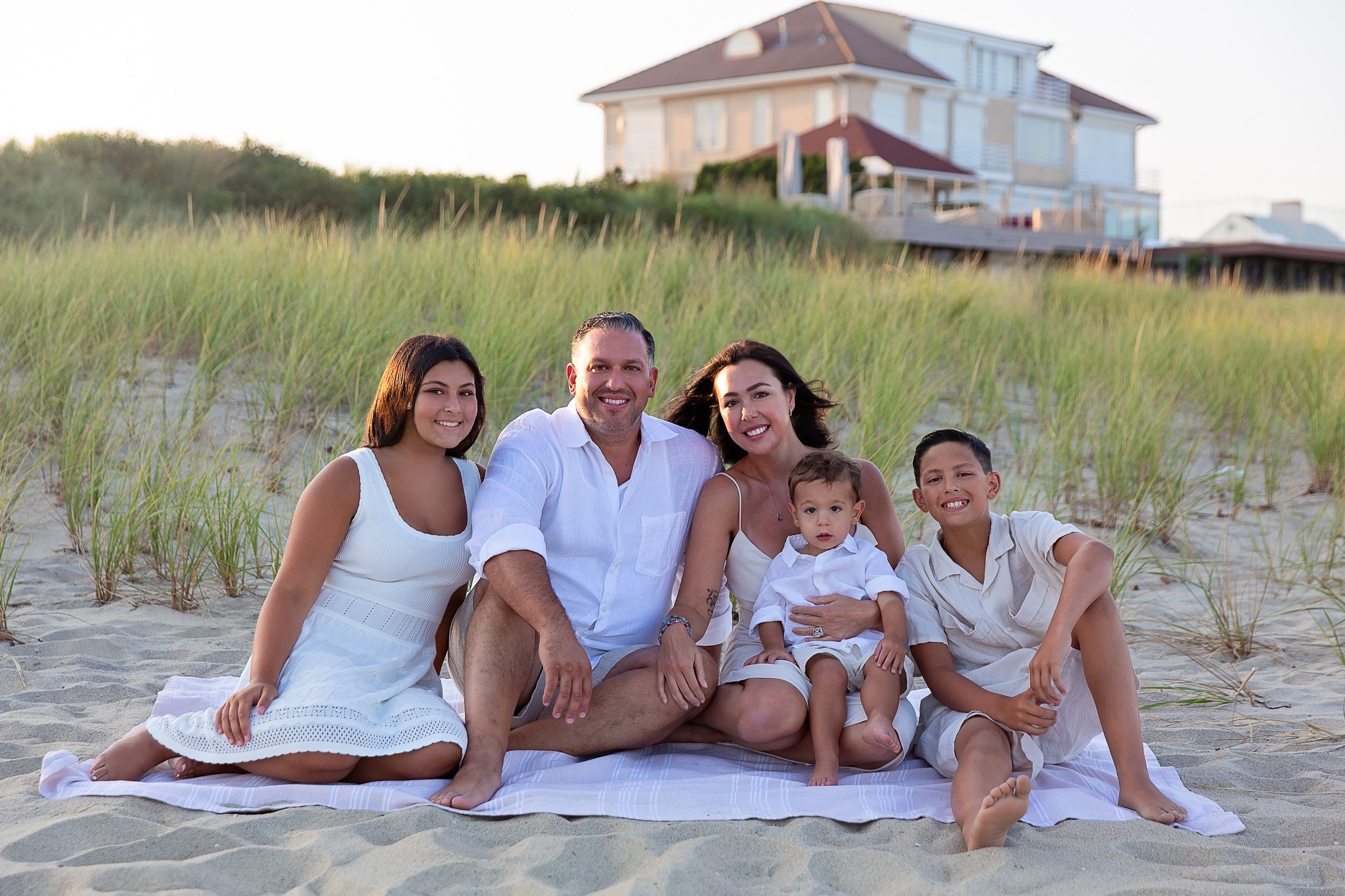 Family of five sitting on a blanket on the beach with a house in the background, smiling and enjoying the day.