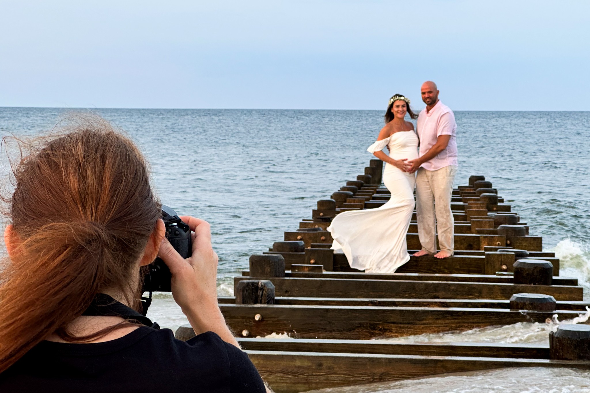 A woman in a white dress and a man in light-colored clothing stand on a wooden pier by the water, being photographed by a person with red hair. The sky is clear.