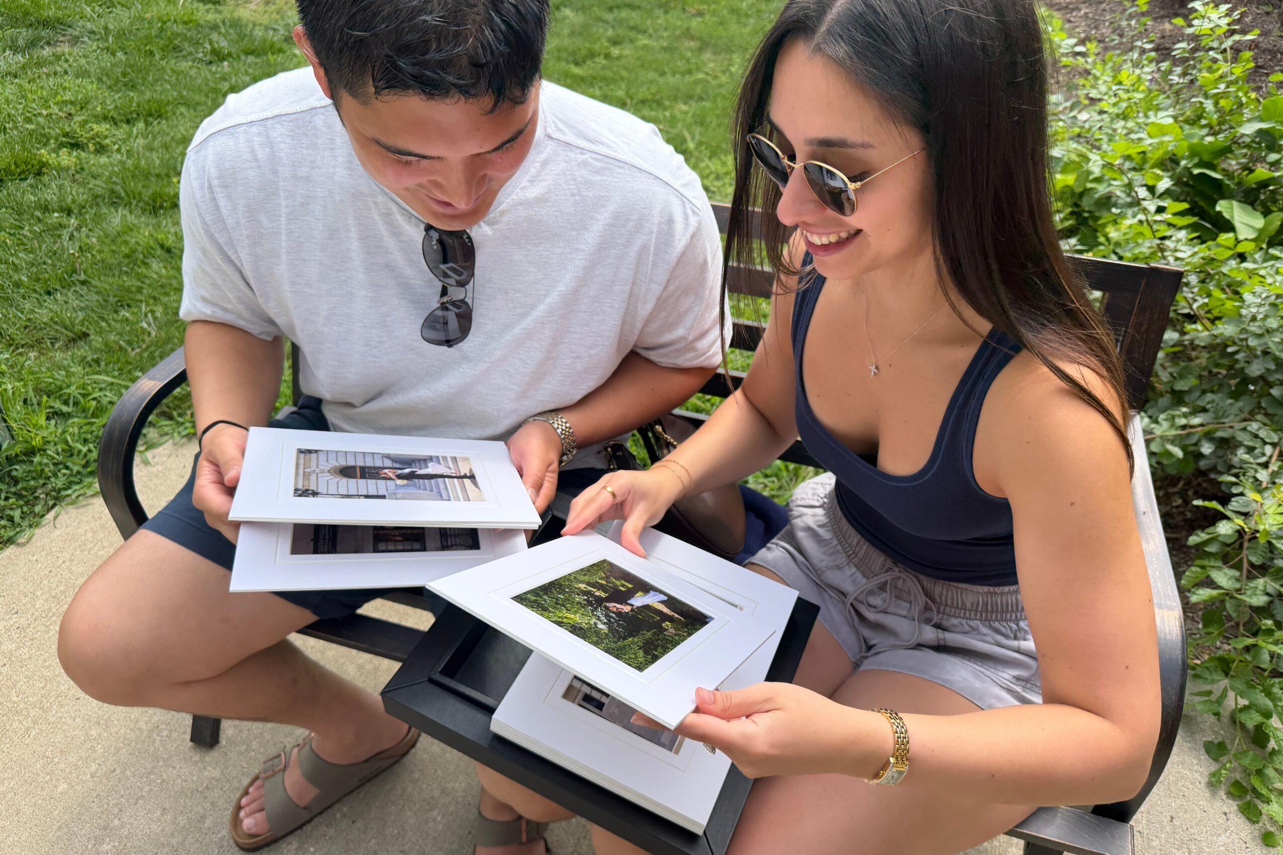 Two people sitting outdoors looking at photographs on a tablet and printed photos, smiling and enjoying the moment.