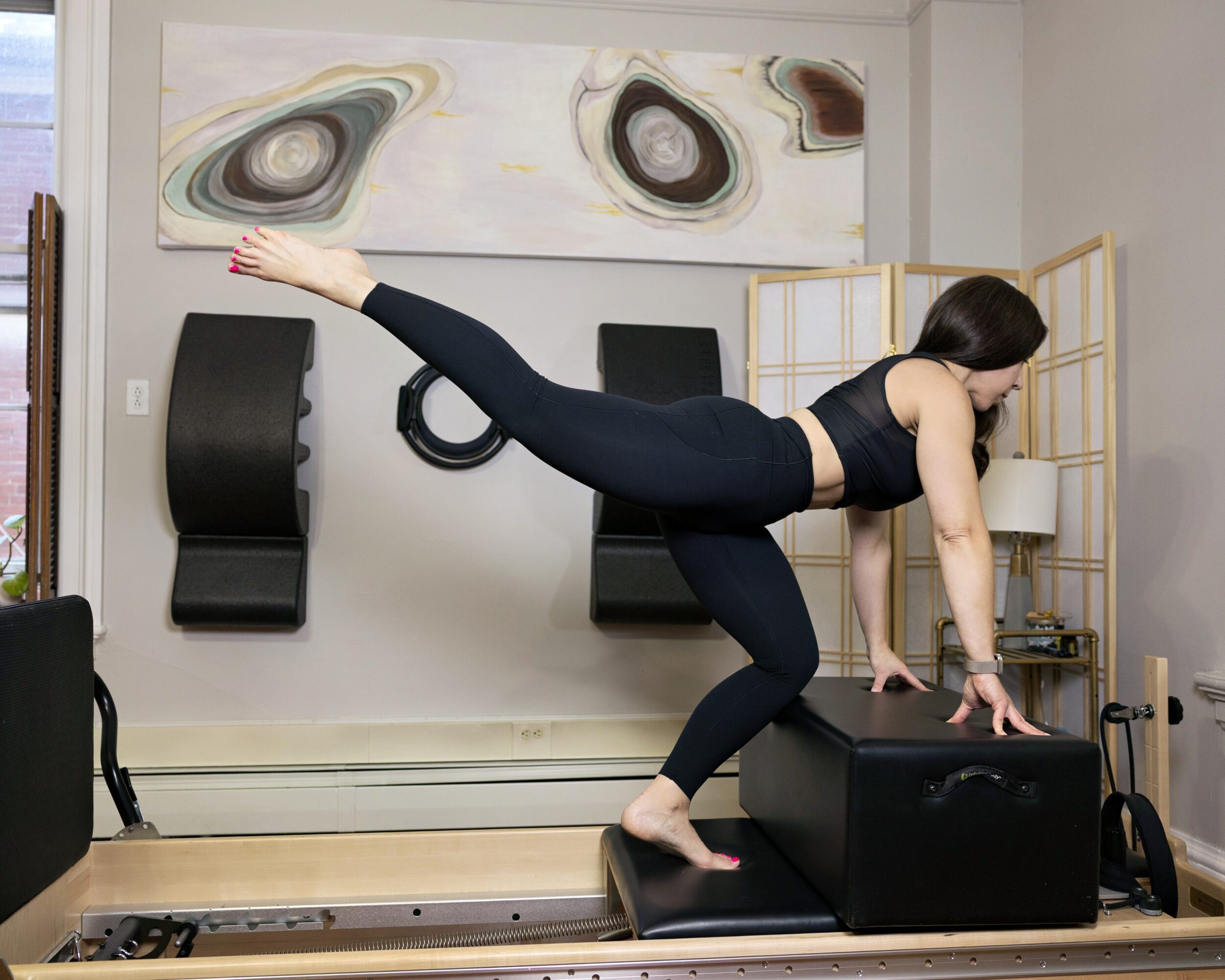 Woman in black workout clothes balances on one leg with the other extended back, hands on a black box, in a fitness studio.