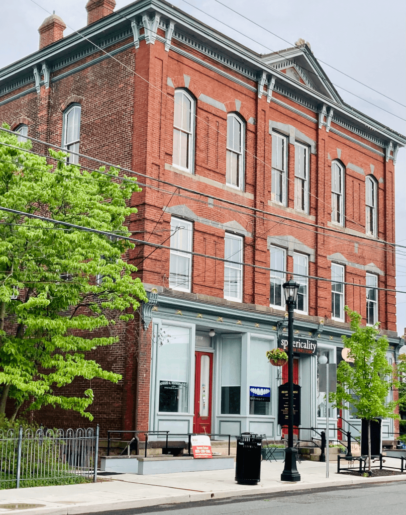 Three-story red brick building with white window frames and a storefront on the ground floor, trees, and street furniture in front.