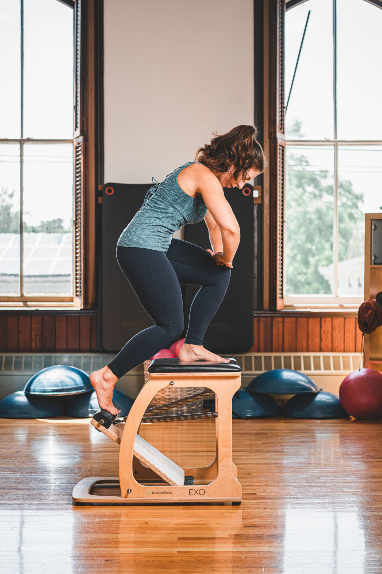 Woman in workout clothes jumps onto a wooden box in a gym with large windows and exercise equipment.