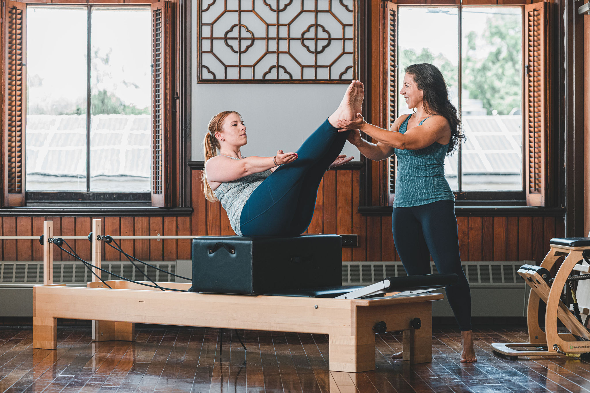 Two women practicing yoga in a room with wooden walls and large windows, one holding the other's feet in a pose.