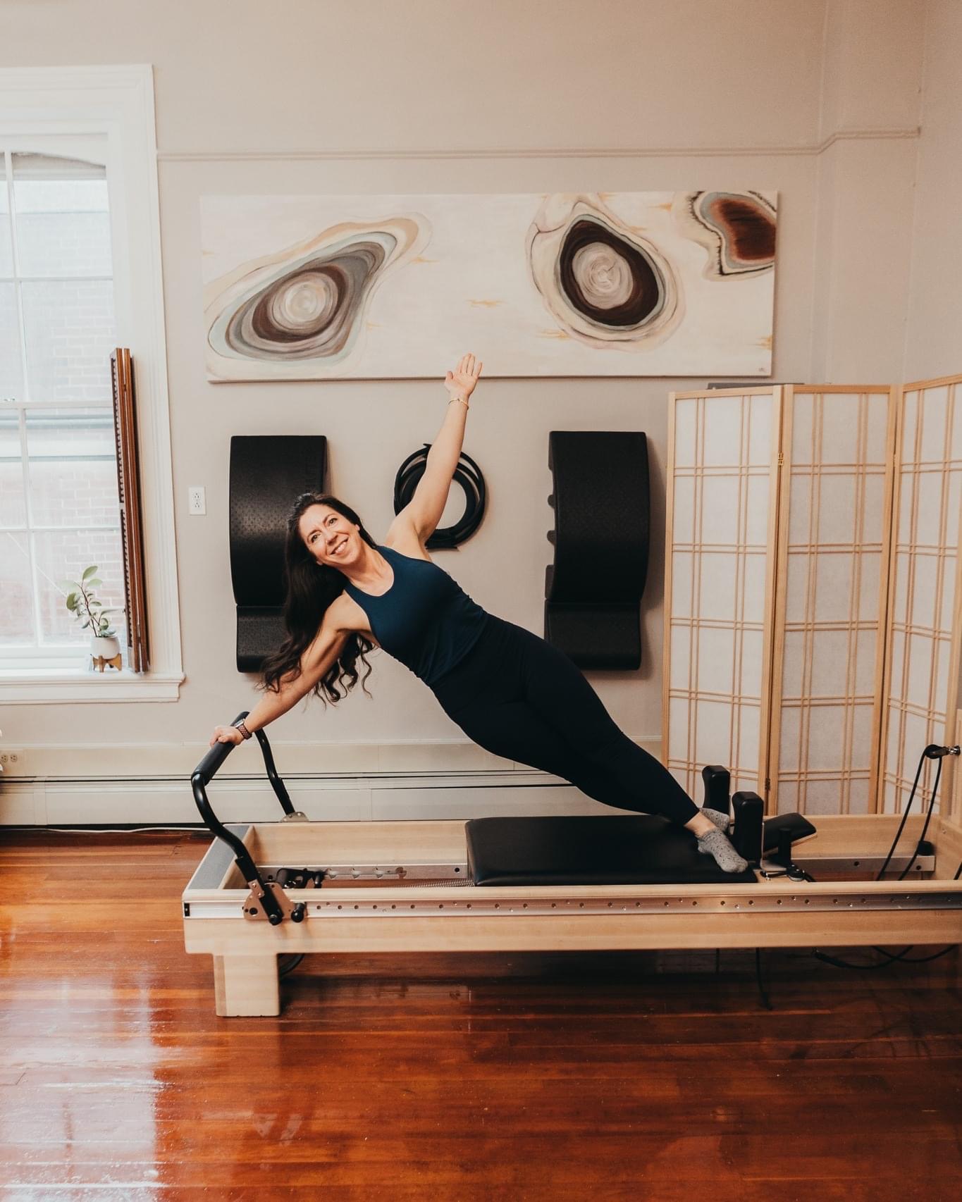 Woman in black workout clothes performs side plank on a Pilates reformer machine in a bright room with artwork and a folding screen.