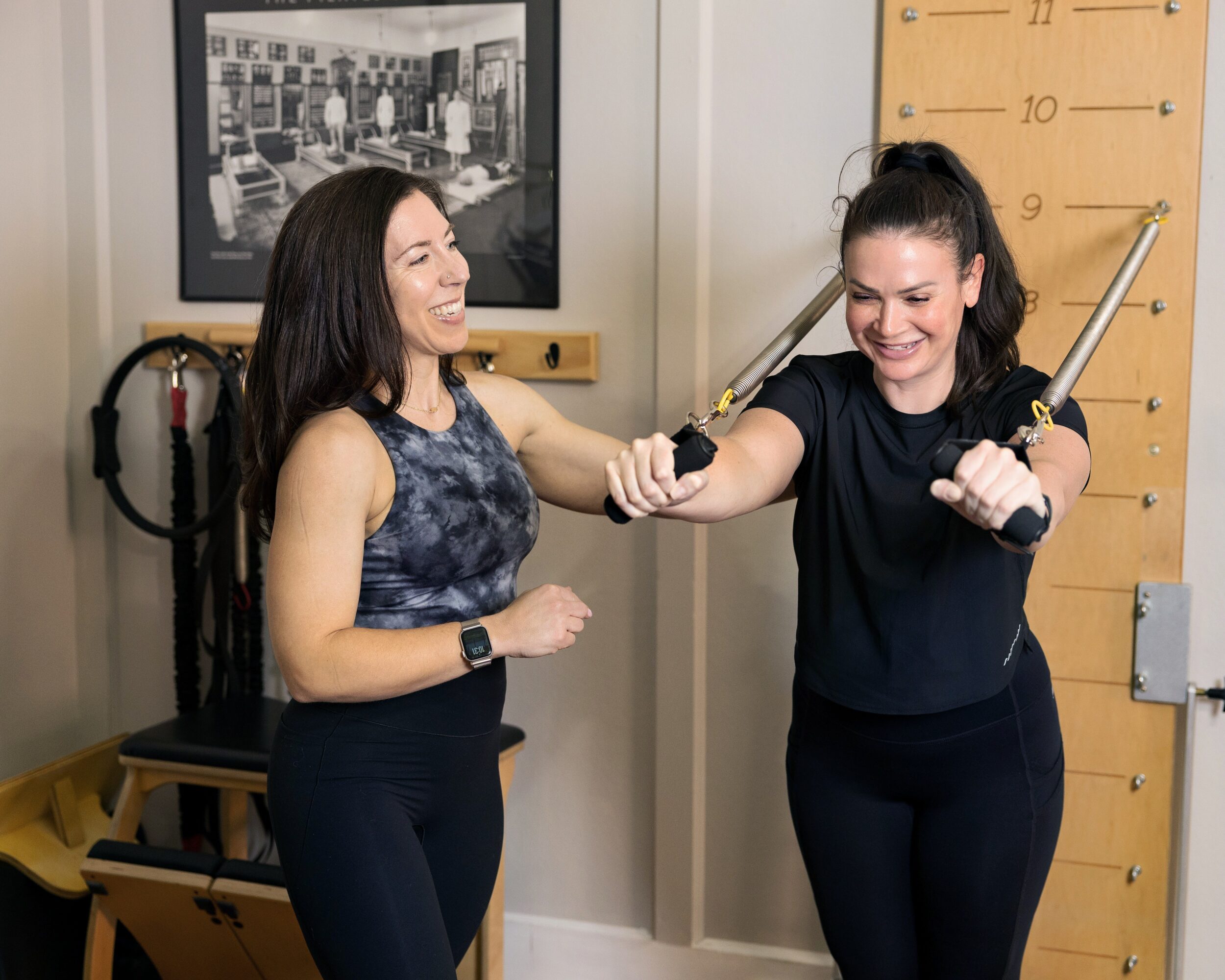 Two women in workout clothes smiling and exercising with resistance bands in a gym, one helping the other.