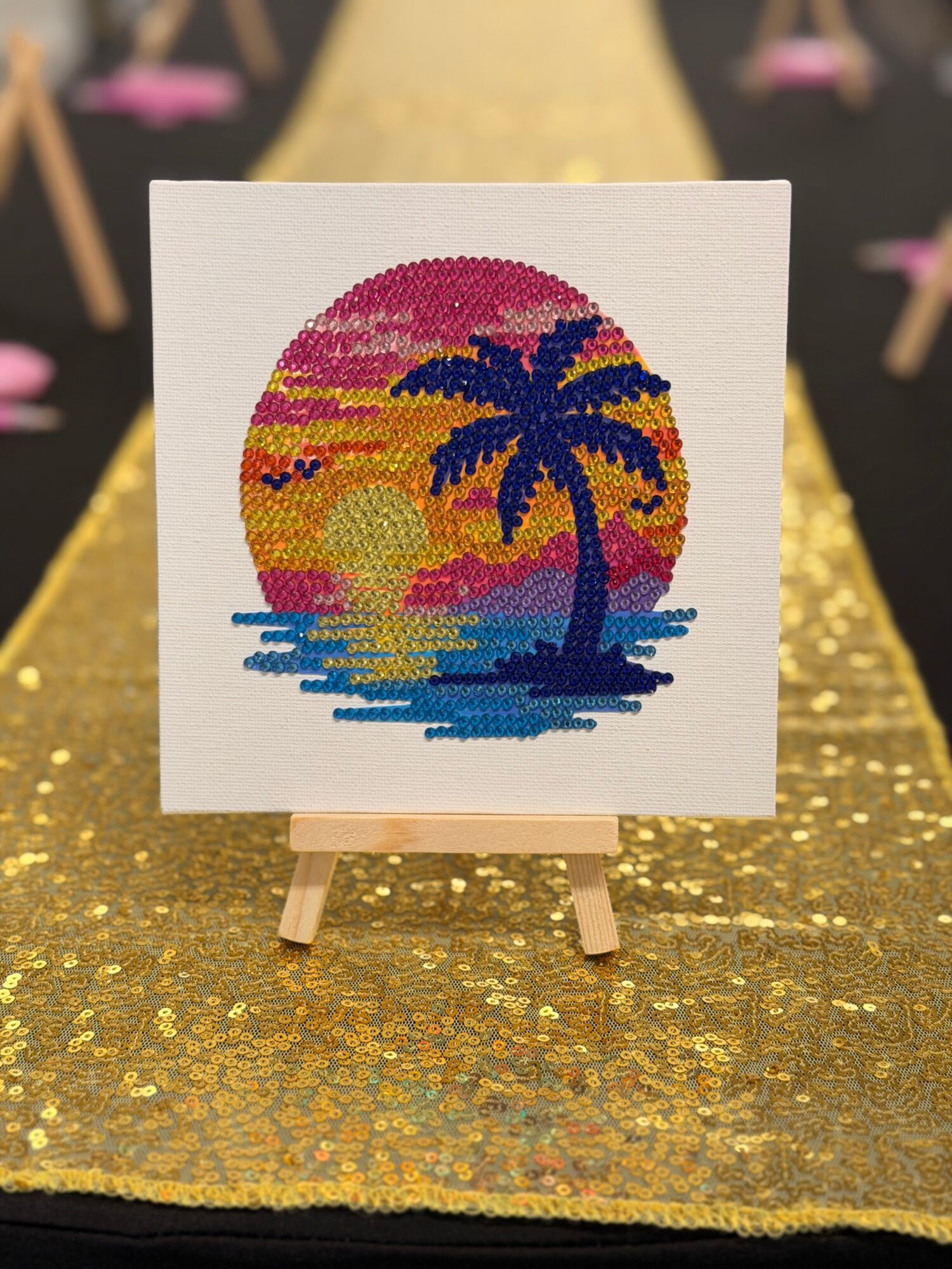 Cross-stitch artwork of a sunset with a palm tree over water, on a small easel, on a gold surface.