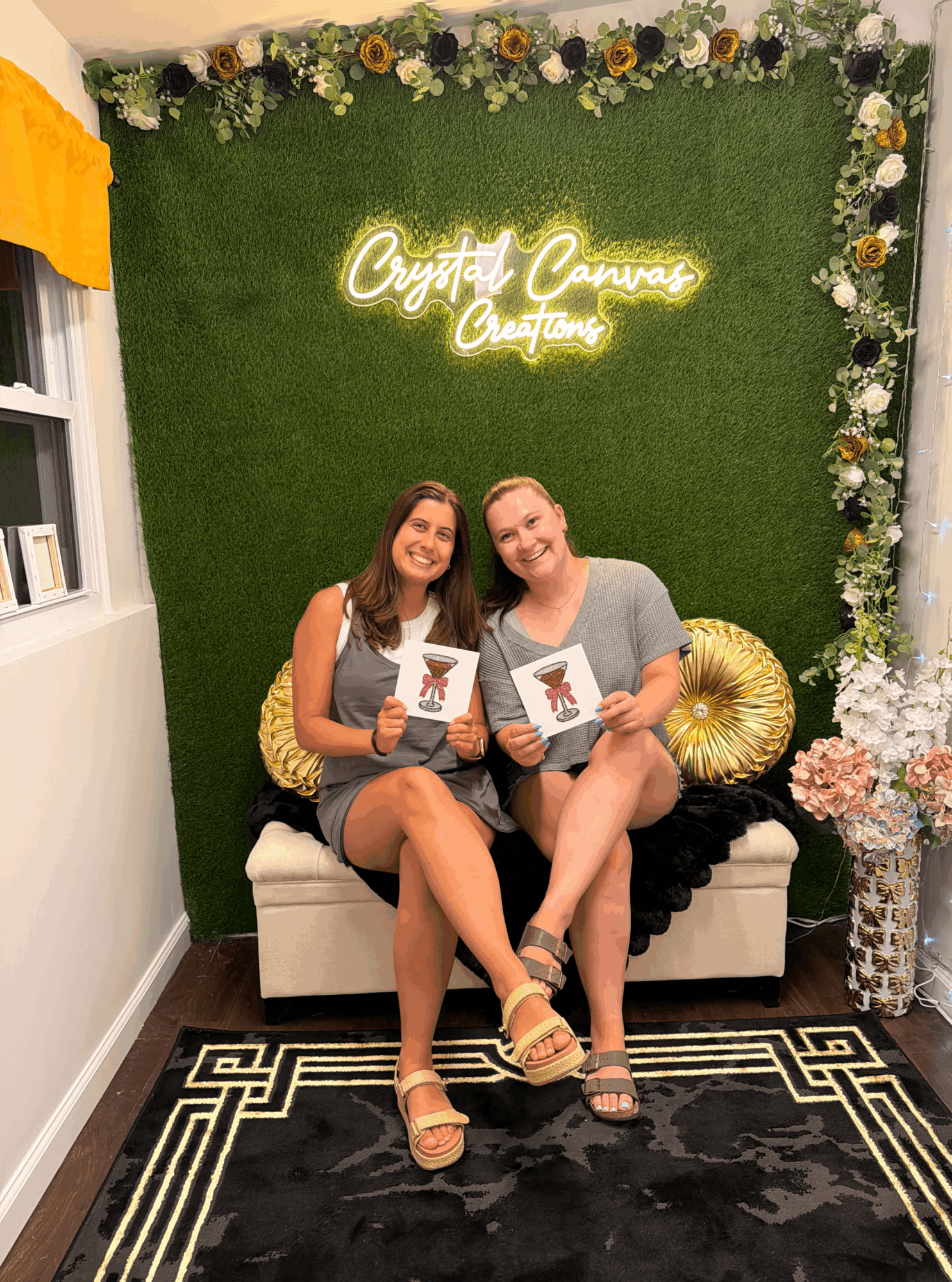 Two women sit on a white sofa with gold pillows, holding books, in front of a green wall with a neon sign and floral decorations.