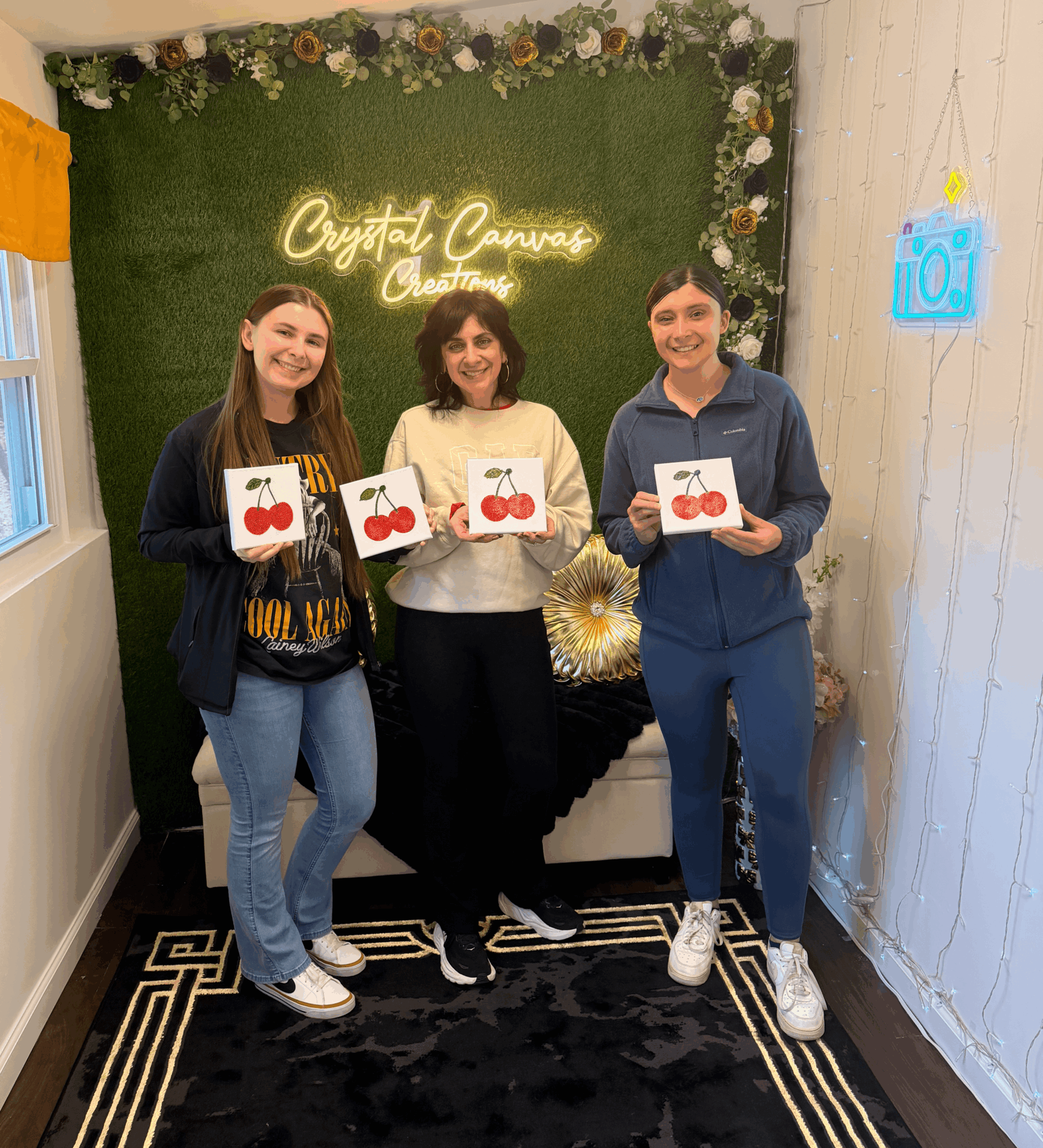 Three people stand indoors holding cards with cherries, smiling, in front of a green wall with a neon sign, decorated with flowers.