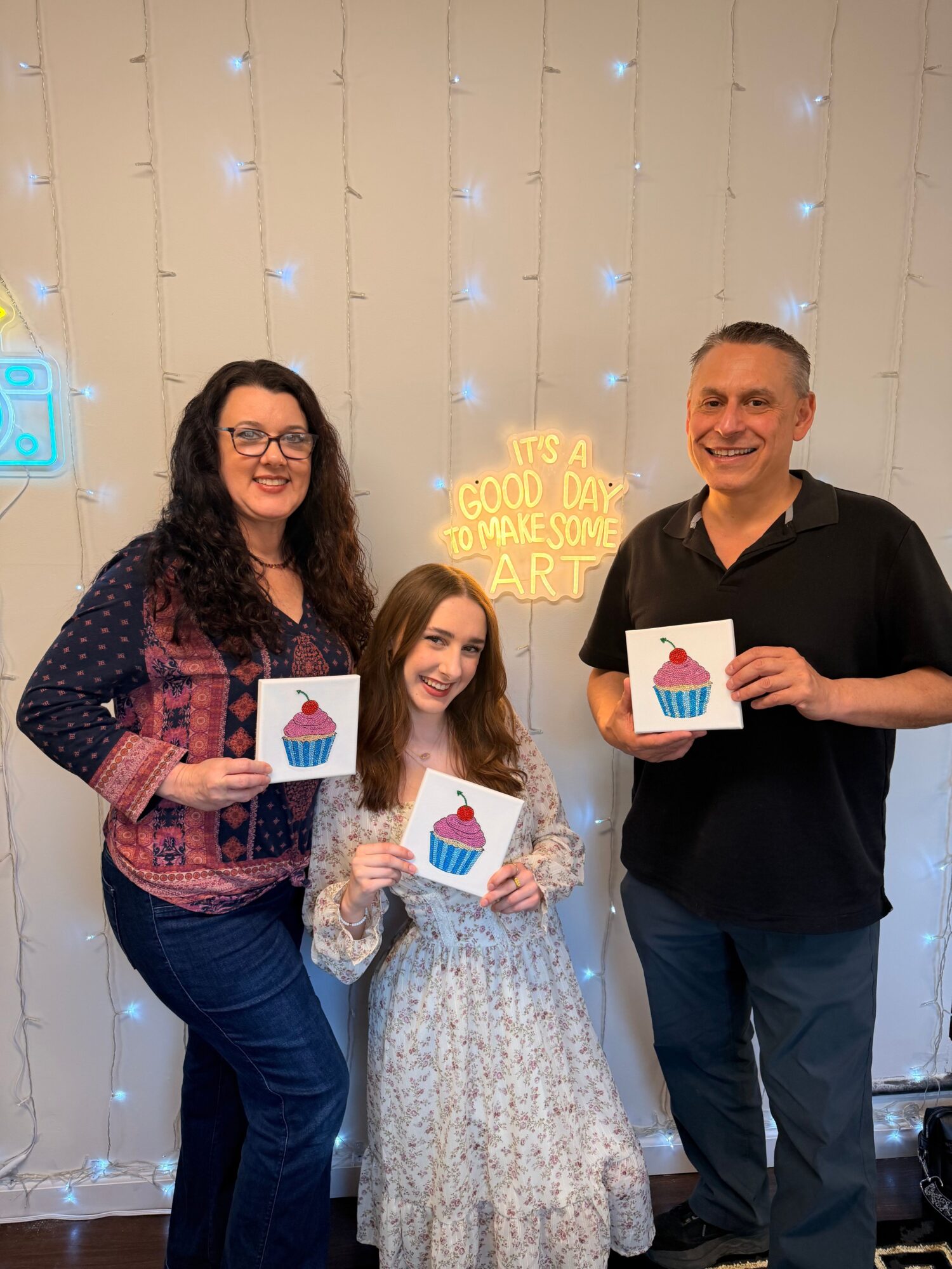 Three people holding certificates, standing in front of a decorated wall with a sign and string lights.