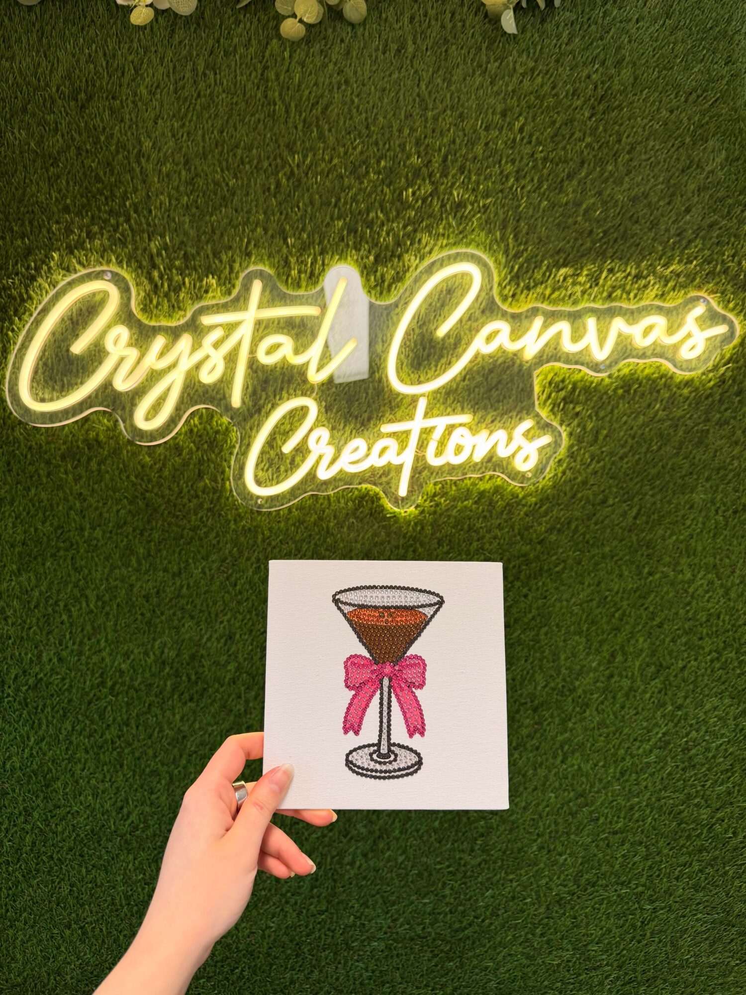 Hand holding a card with a drawing of a glass with a pink drink and a straw, on green grass background.