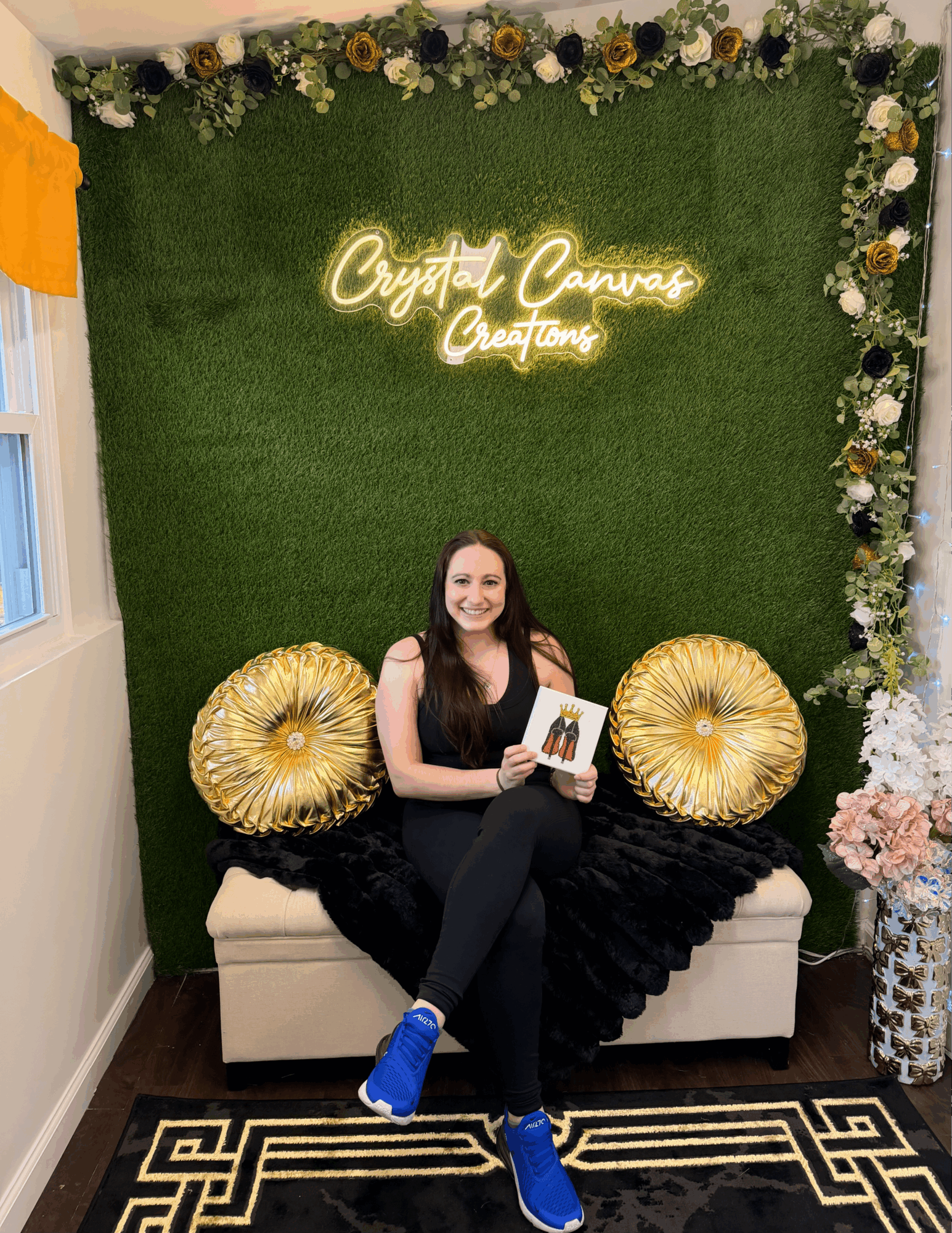 Woman sitting on a sofa holding a book, with a green wall, floral decorations, and neon sign behind her.
