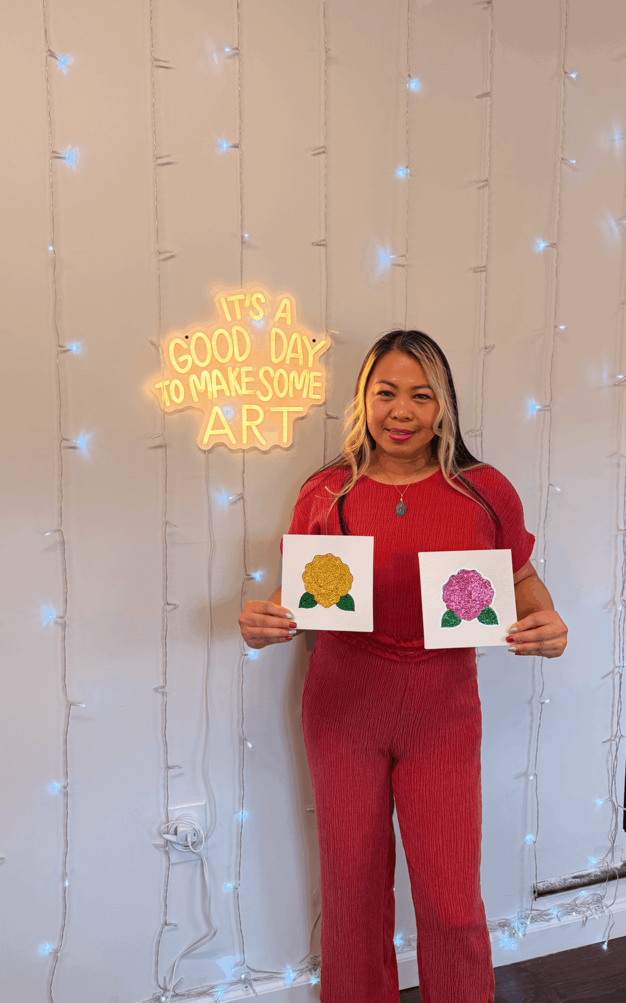 Woman in red holding two paintings of flowers, standing in front of a wall with string lights and a neon sign.