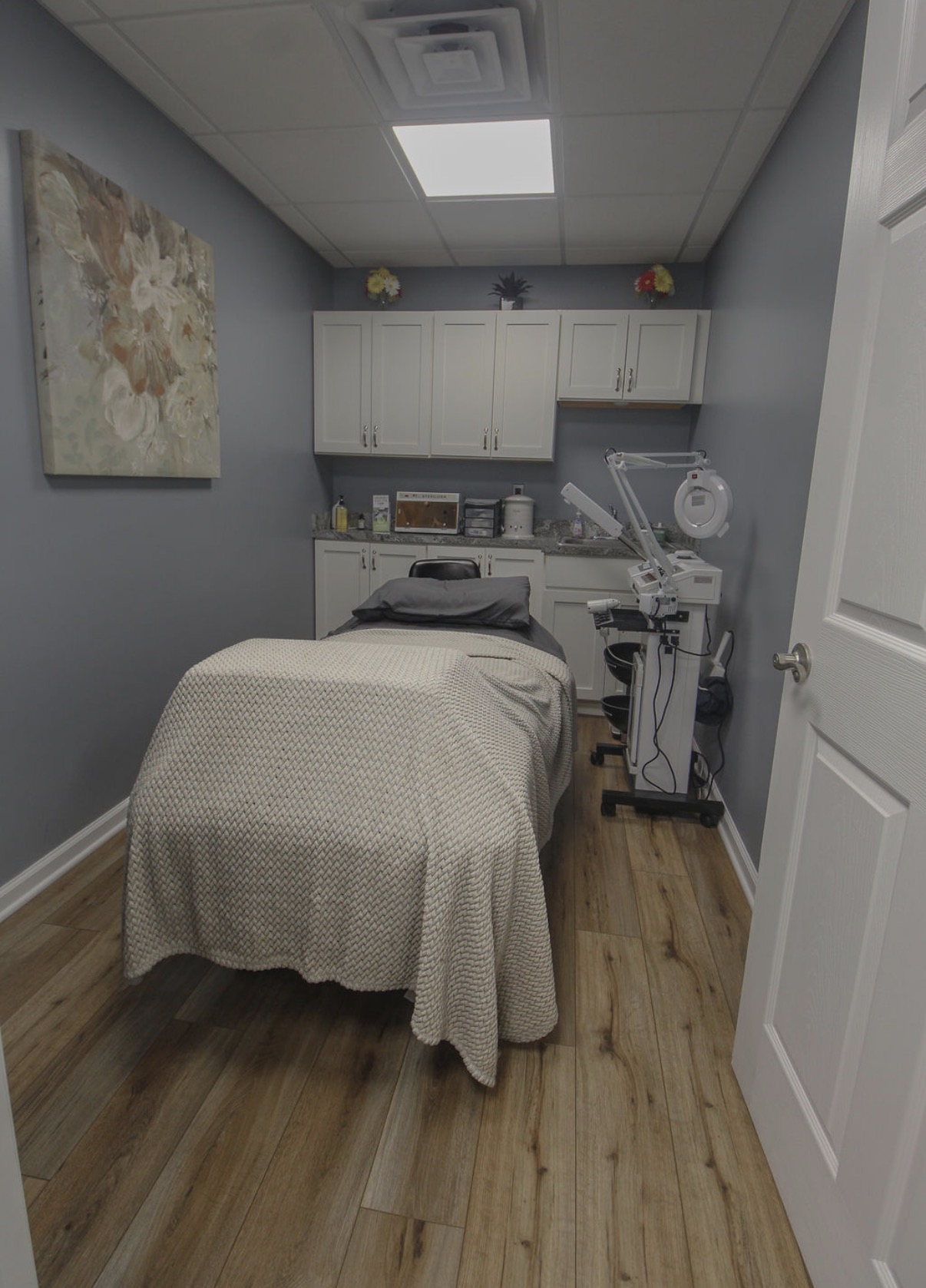 Medical examination room with a bed, cabinets, and medical equipment, walls painted light blue, wooden floor, ceiling light.