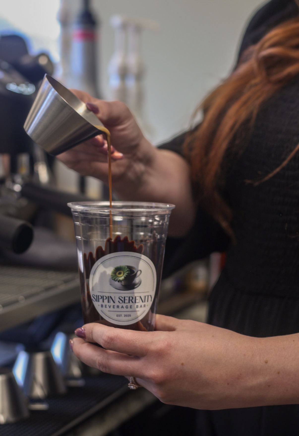 Person pours coffee into a clear cup with a logo, in a setting with coffee equipment in the background.