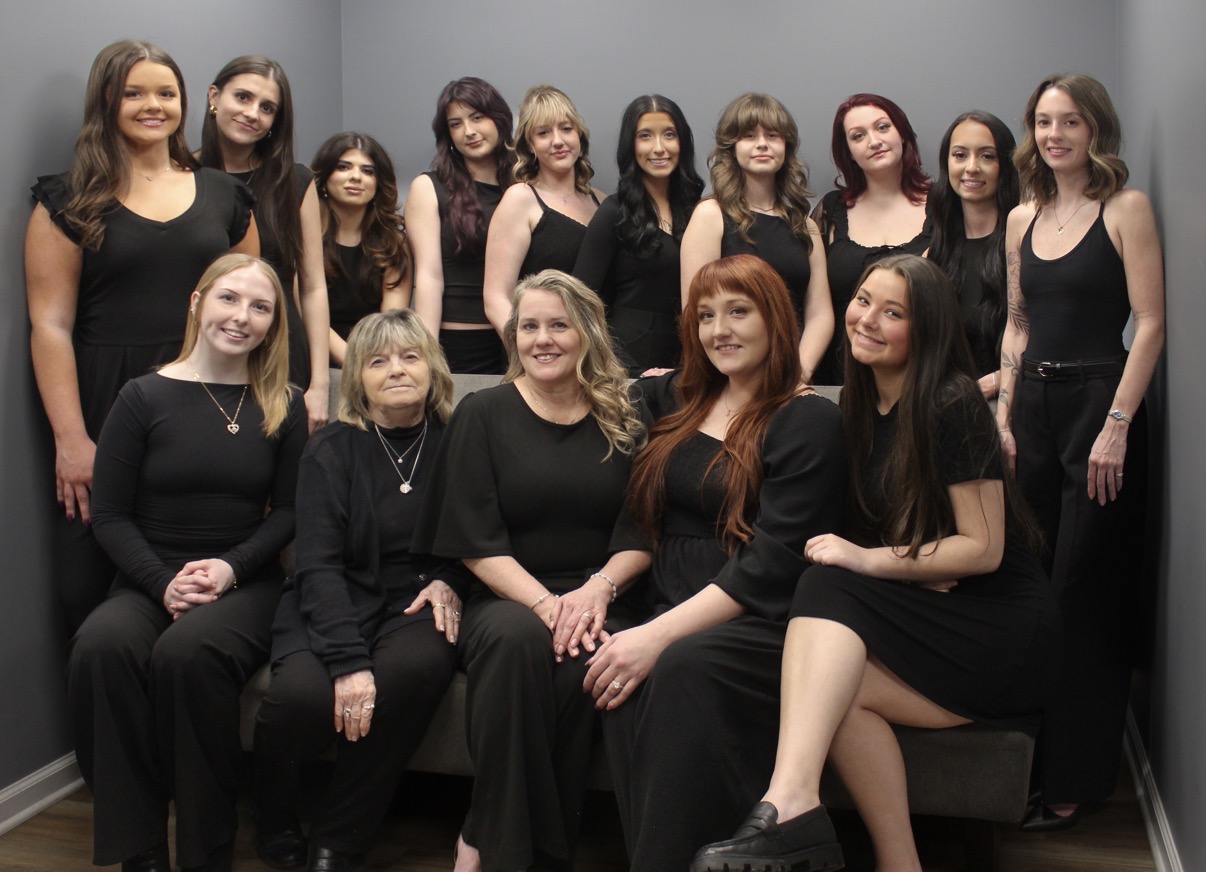 Group of fifteen women in black dresses posing together indoors against a gray wall.