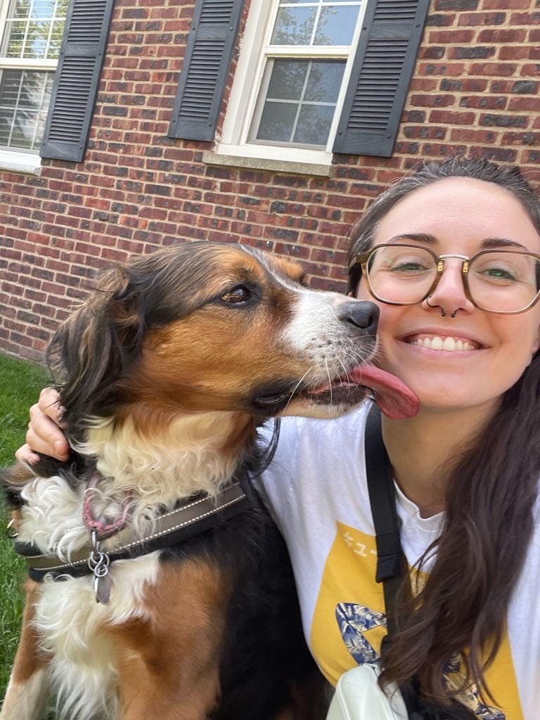 Woman smiling with glasses and a dog outdoors in front of a brick house, woman holding the dog close.