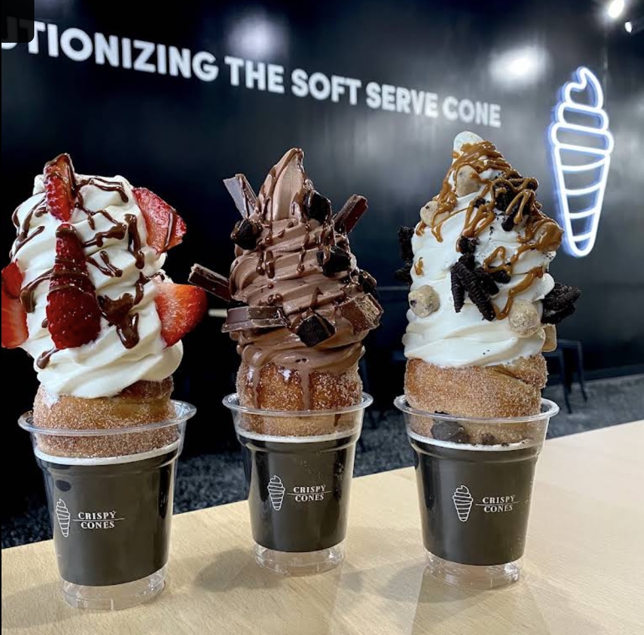 Three soft serve ice cream cones with toppings on a table, with a sign and ice cream graphic in the background.