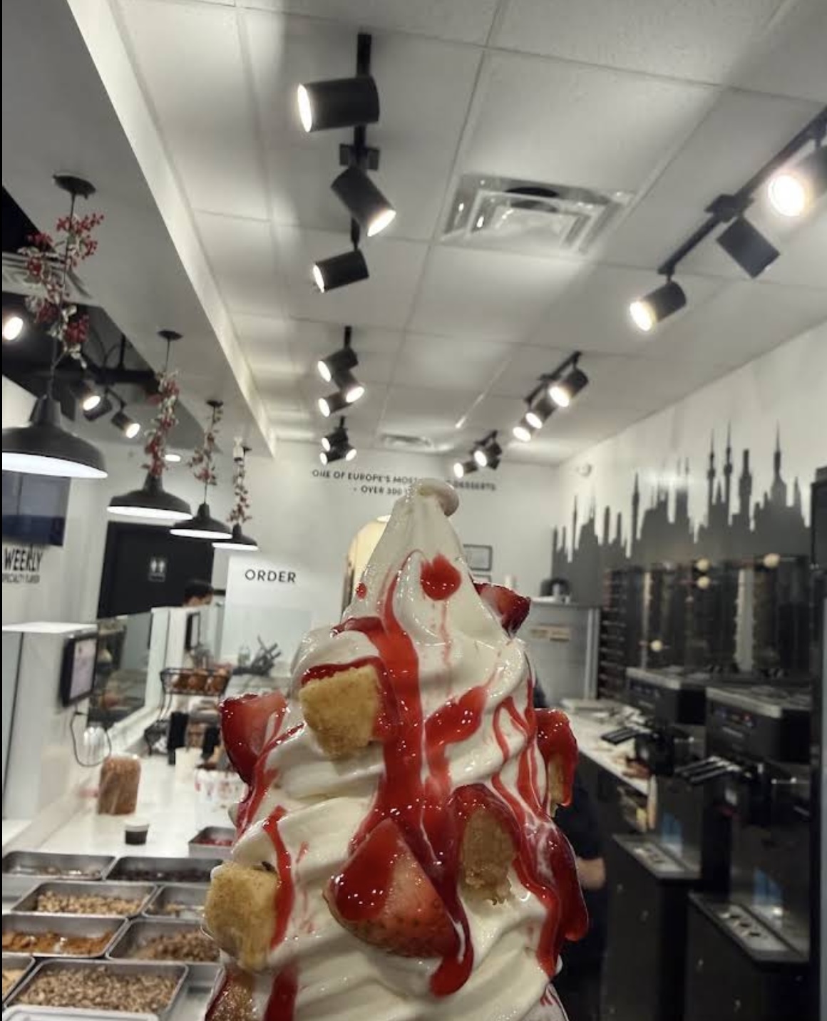 A dessert with whipped cream, strawberries, and sauce on a table in a cafe, with a city skyline mural on the wall.