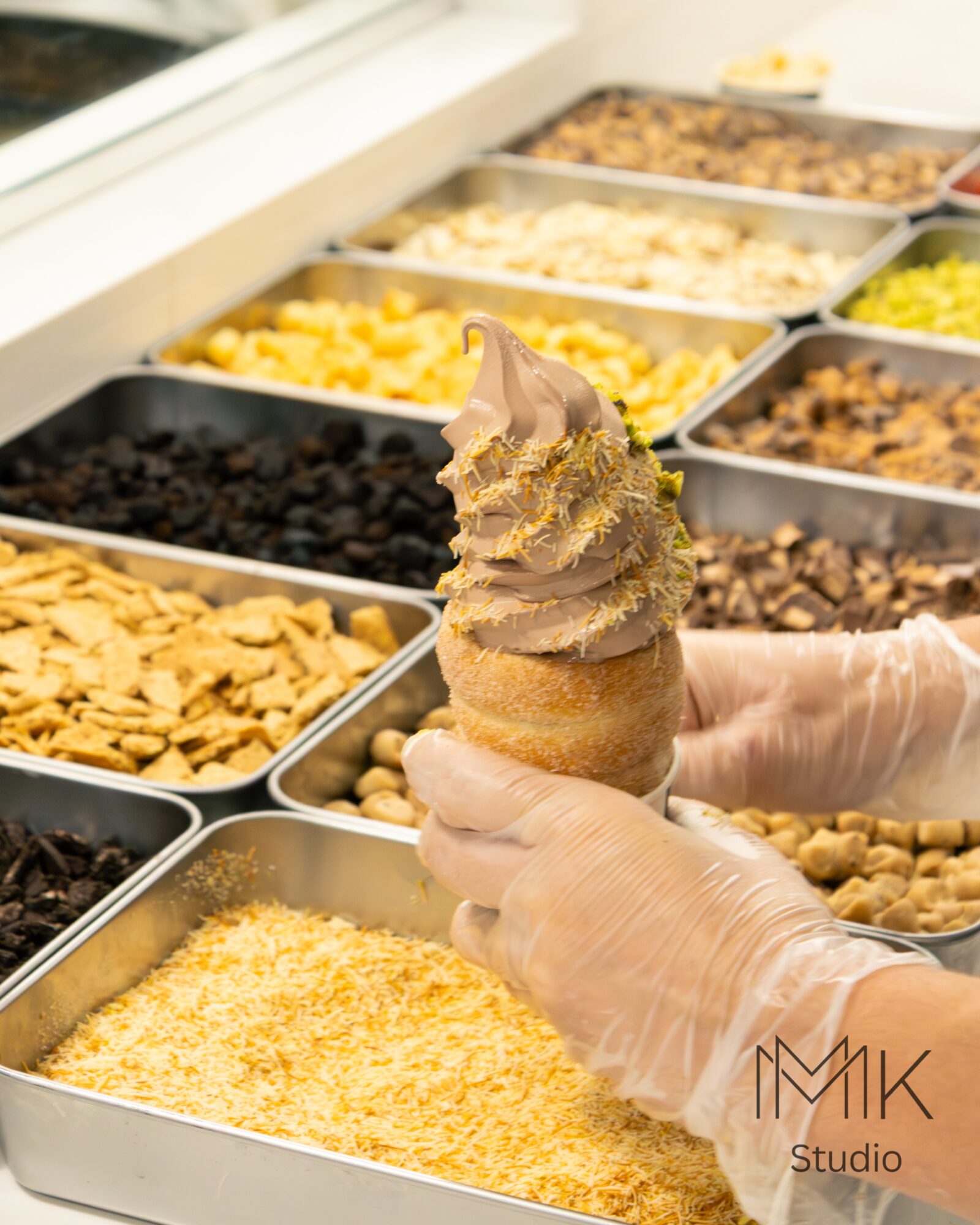 Person wearing gloves holds a soft serve ice cream cone with chocolate and vanilla swirls in front of toppings display.