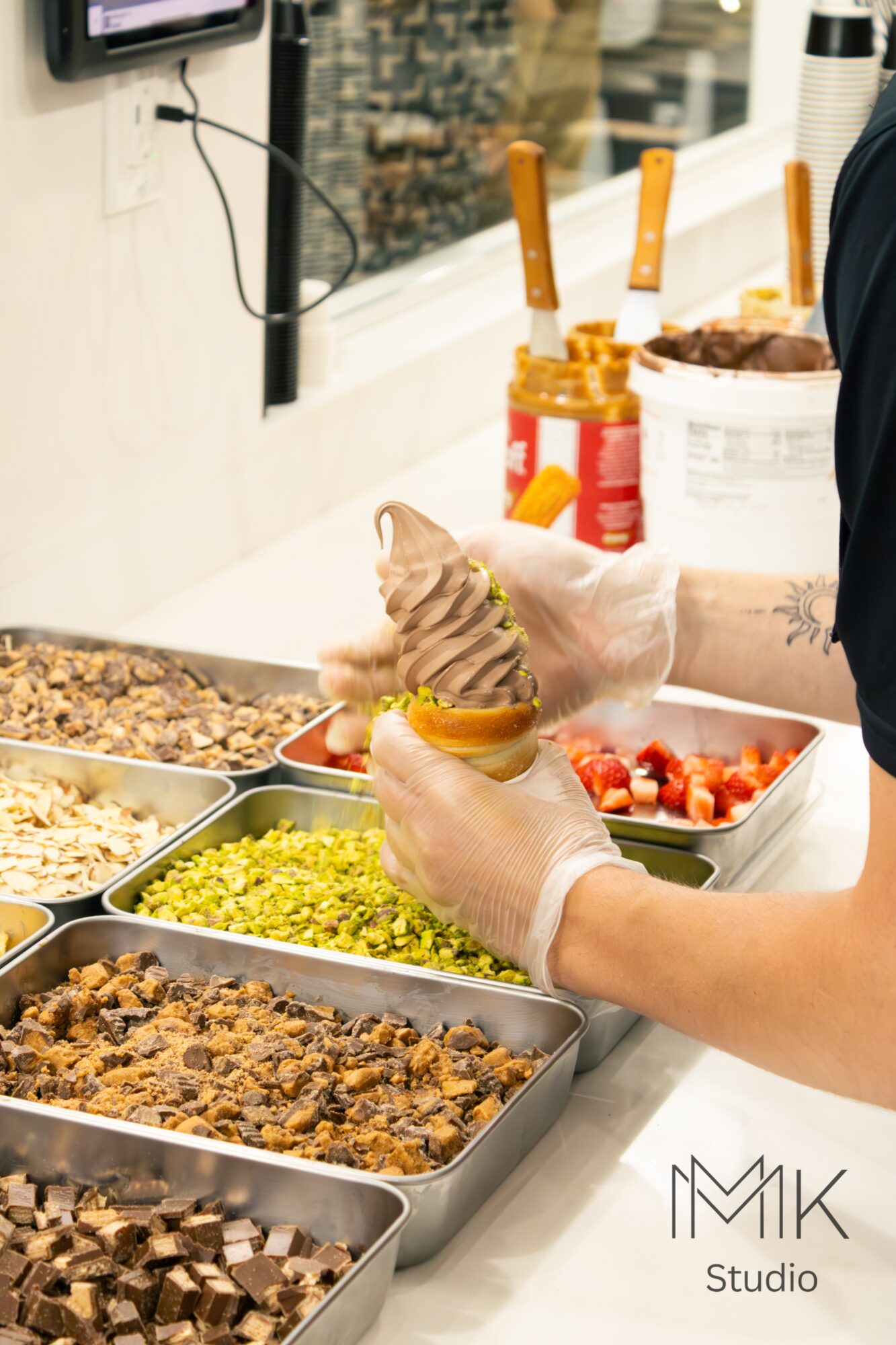Person serving ice cream into a bowl with toppings, surrounded by various food containers, in a bright setting.