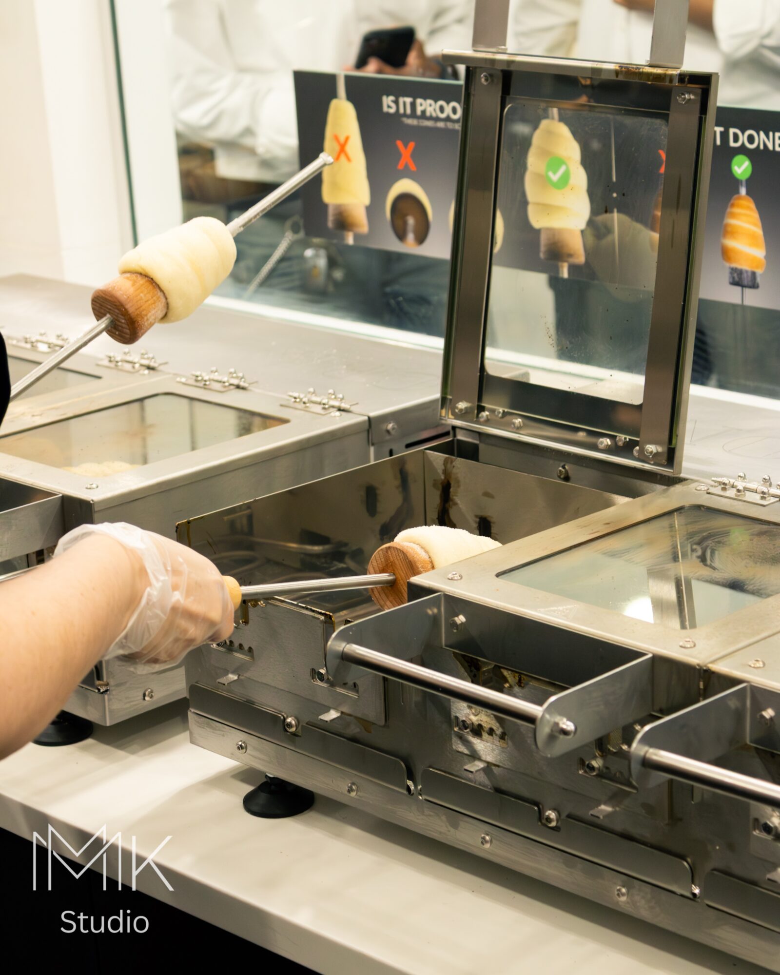 Person using tongs to place ice cream cone into machine, with display screen showing ice cream options, in a shop setting.
