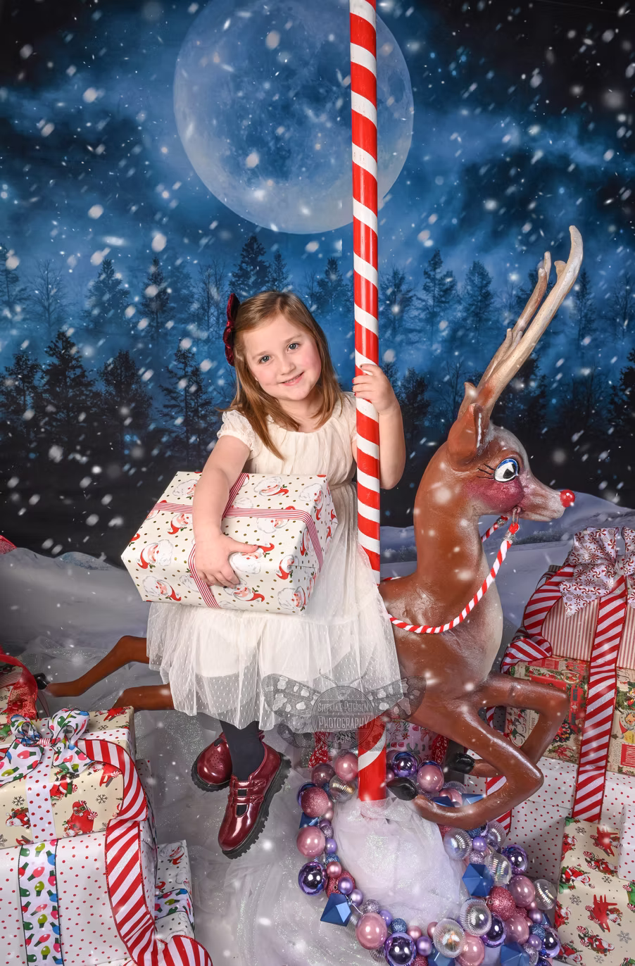 Young girl in a white dress sitting on a carousel reindeer with a snowy night background and moon, holding a gift box.