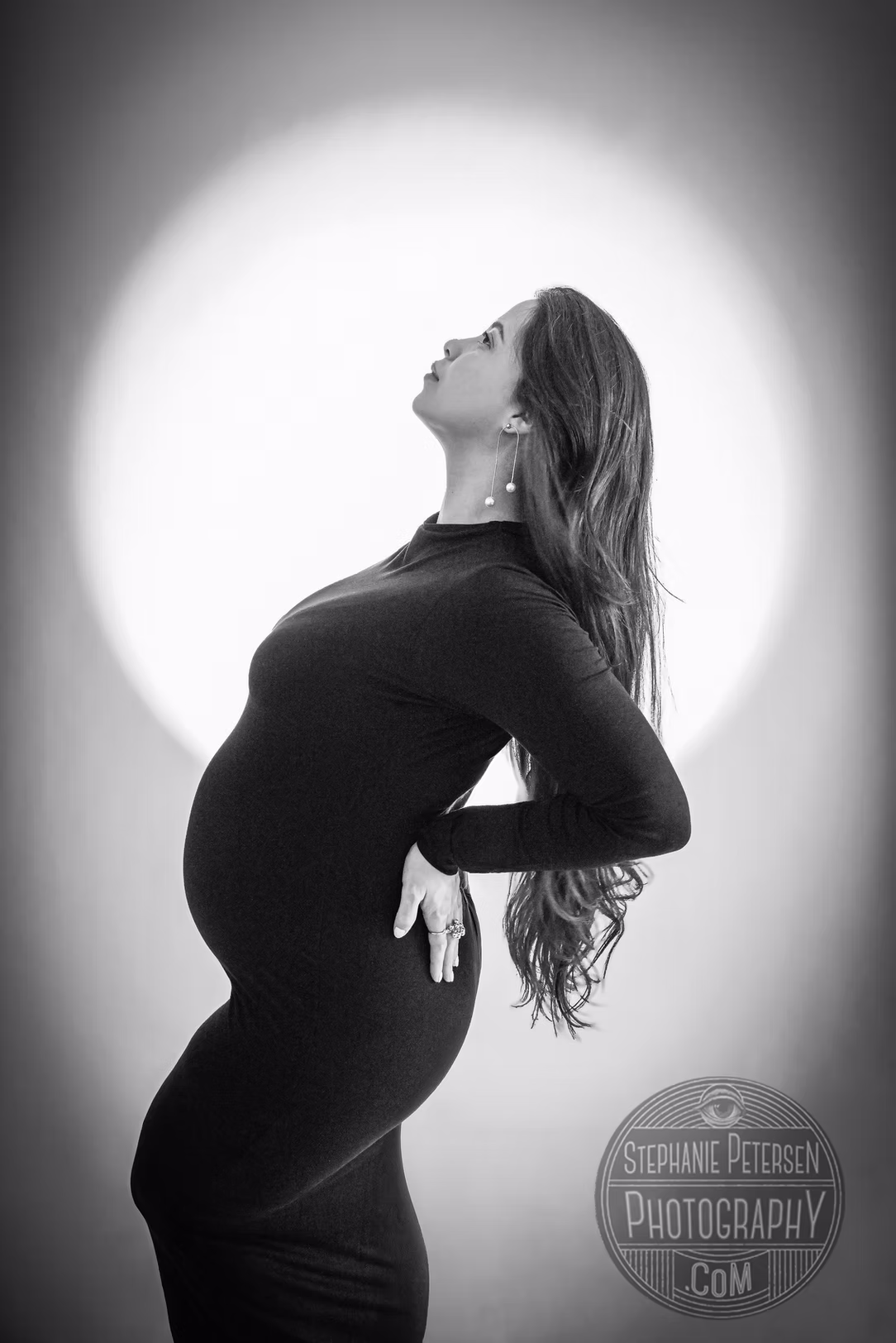 Pregnant woman in a black dress with long hair, standing sideways with her head tilted back, in front of a bright circular background.
