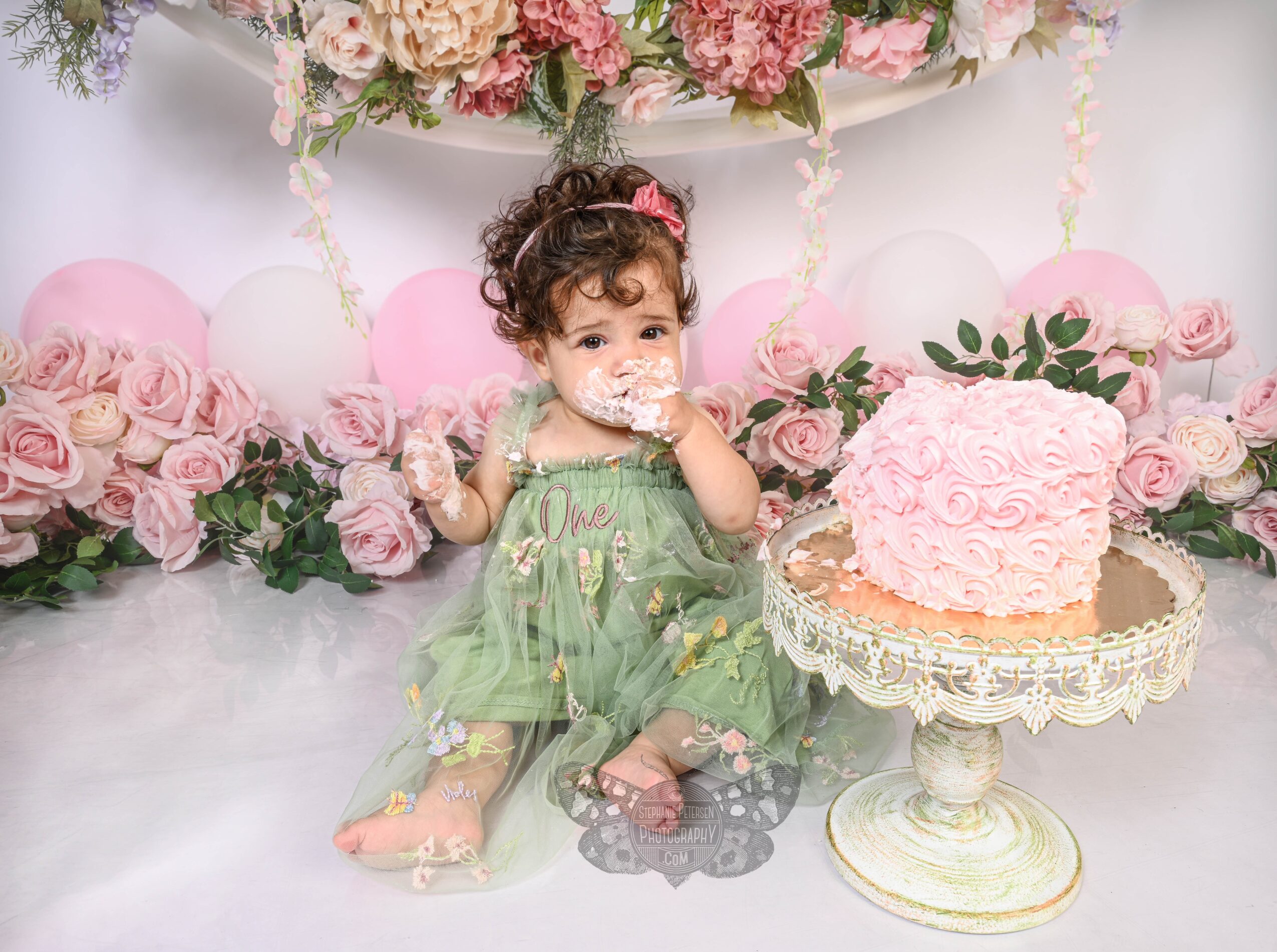 Young girl with curly hair in a green dress sitting on the floor near a pink cake, surrounded by pink and white flowers.