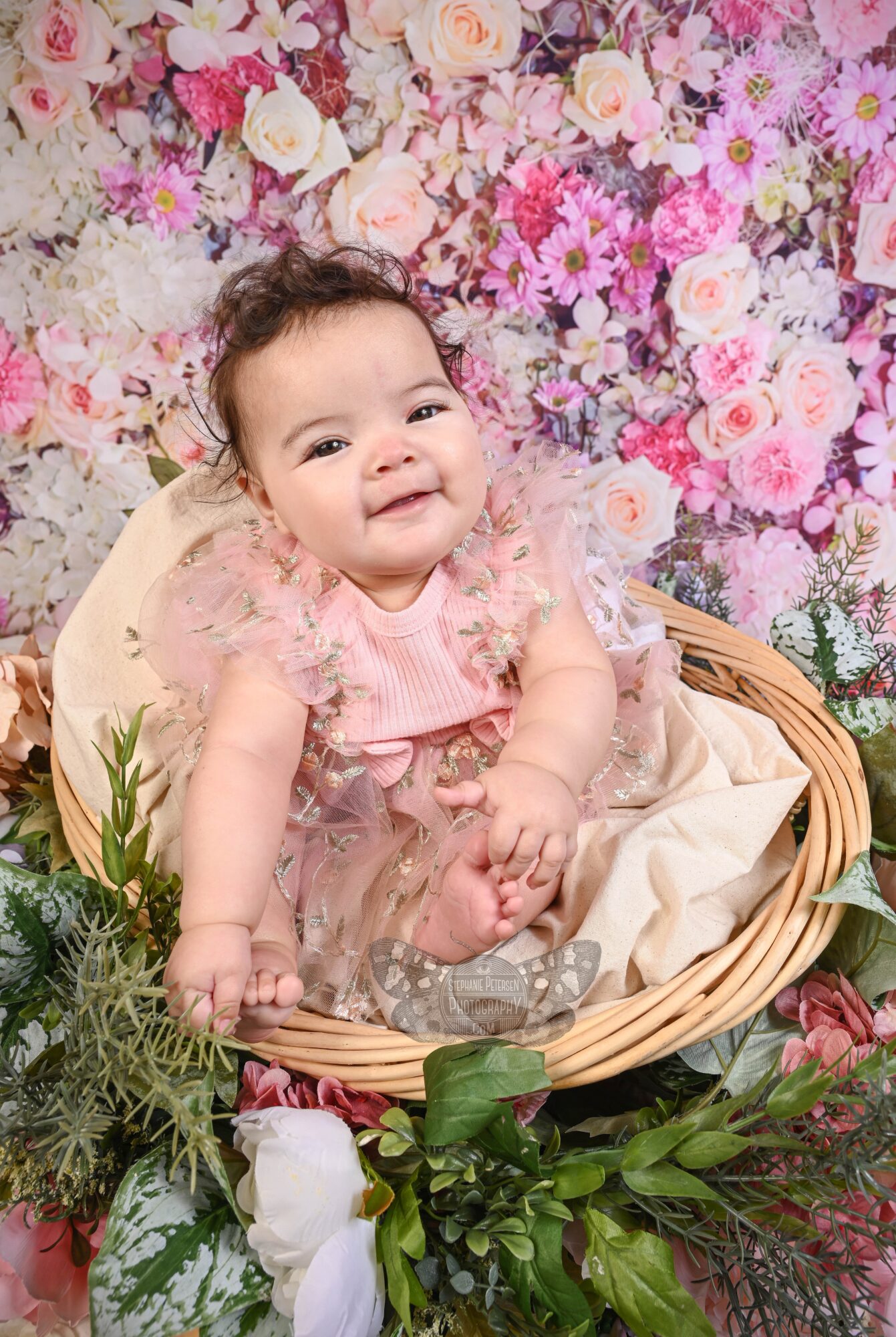 Smiling baby girl sitting in a basket with a floral background, surrounded by greenery and flowers.