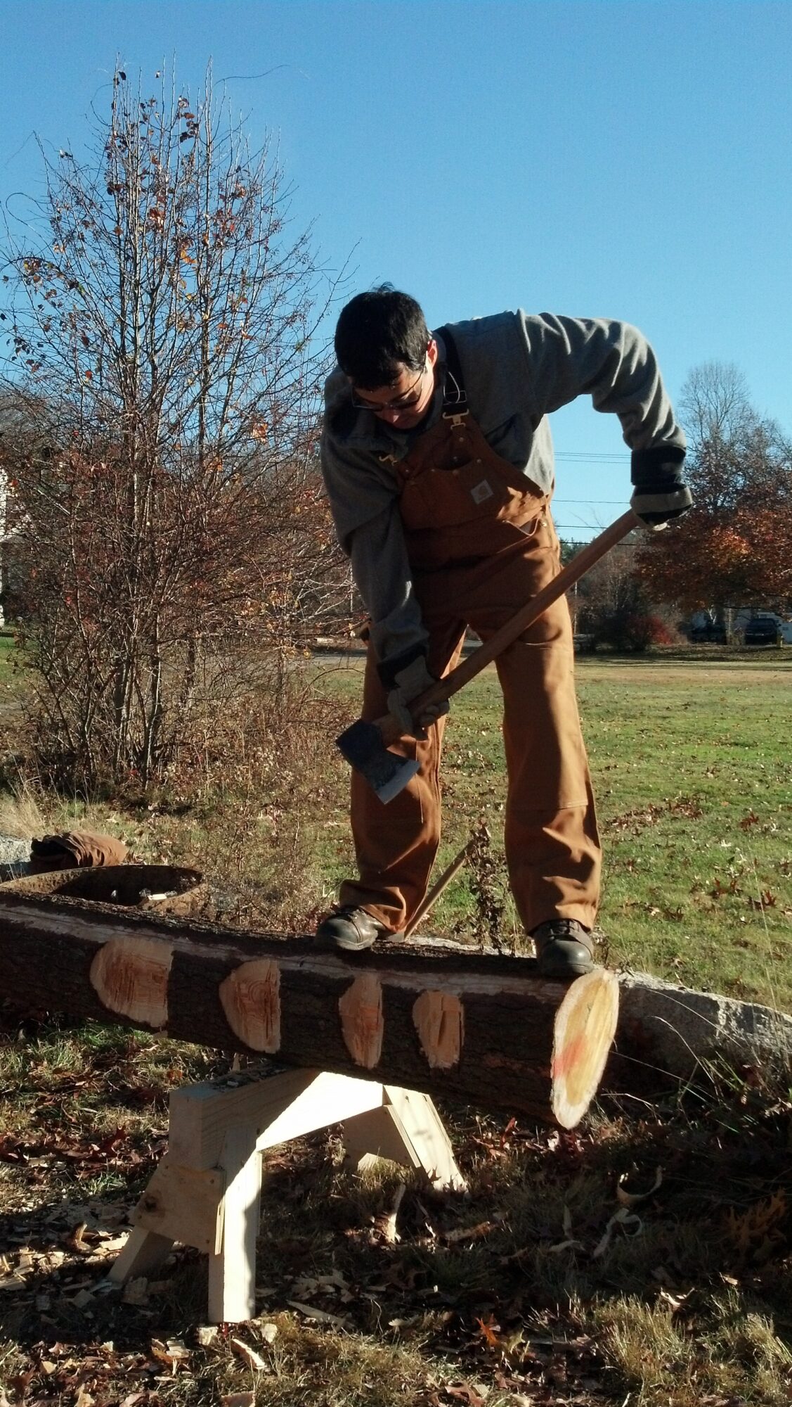 Person balancing on a fallen tree trunk outdoors, wearing brown overalls and a gray jacket, with trees and grass in background.