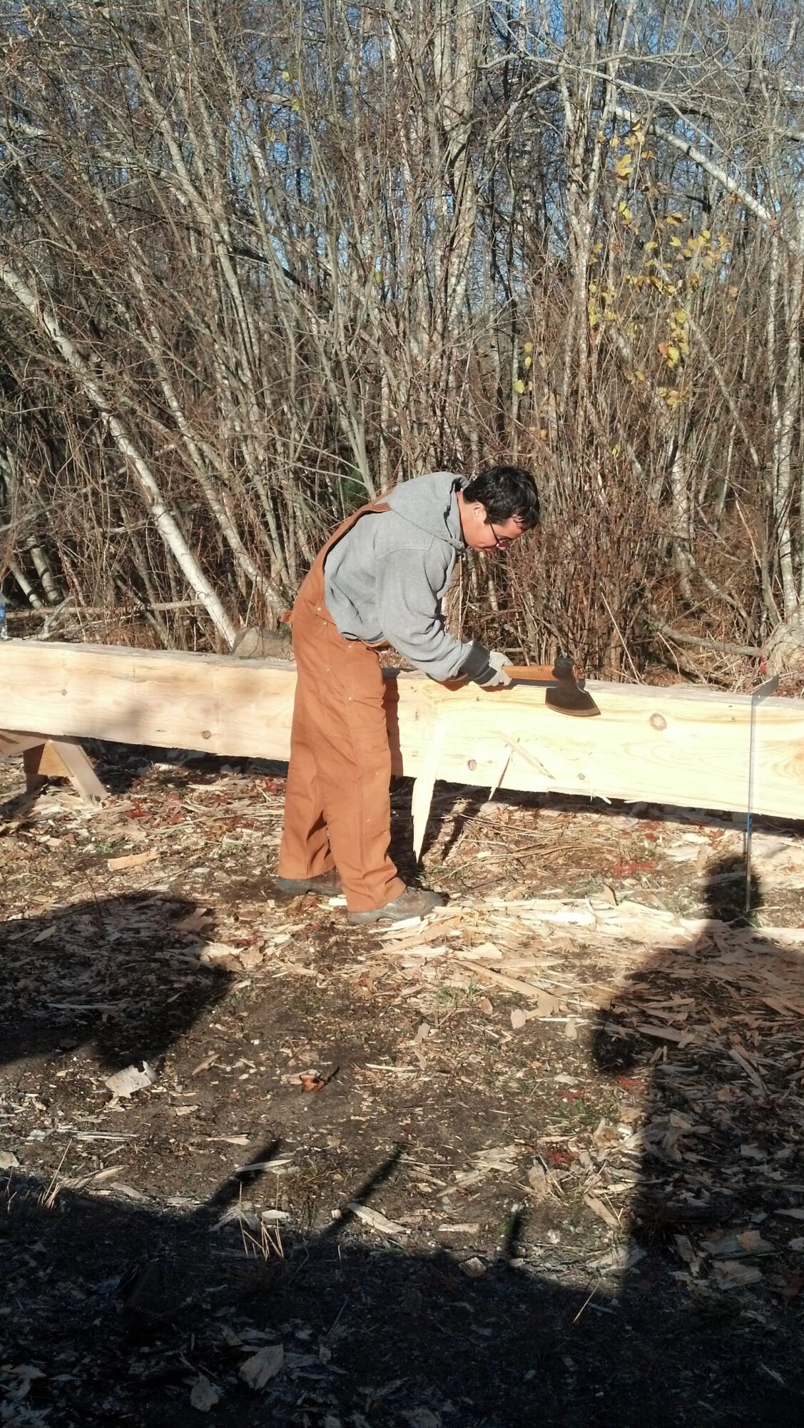 Person working outdoors on a wooden beam, wearing a gray hoodie and brown pants, with trees in the background.