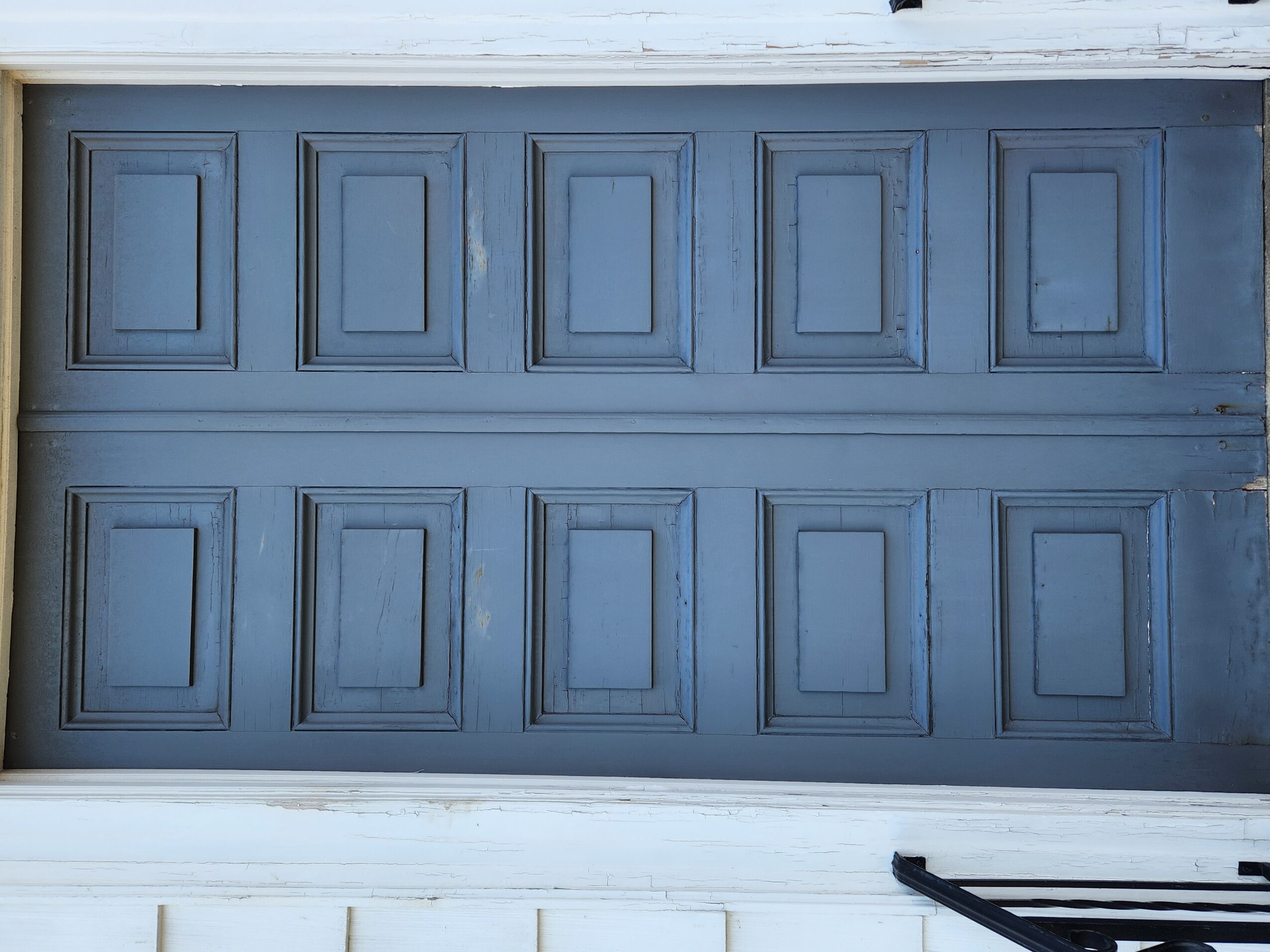 Blue garage door with rectangular panels, two rows, white trim at top and bottom, and a black object at bottom right.