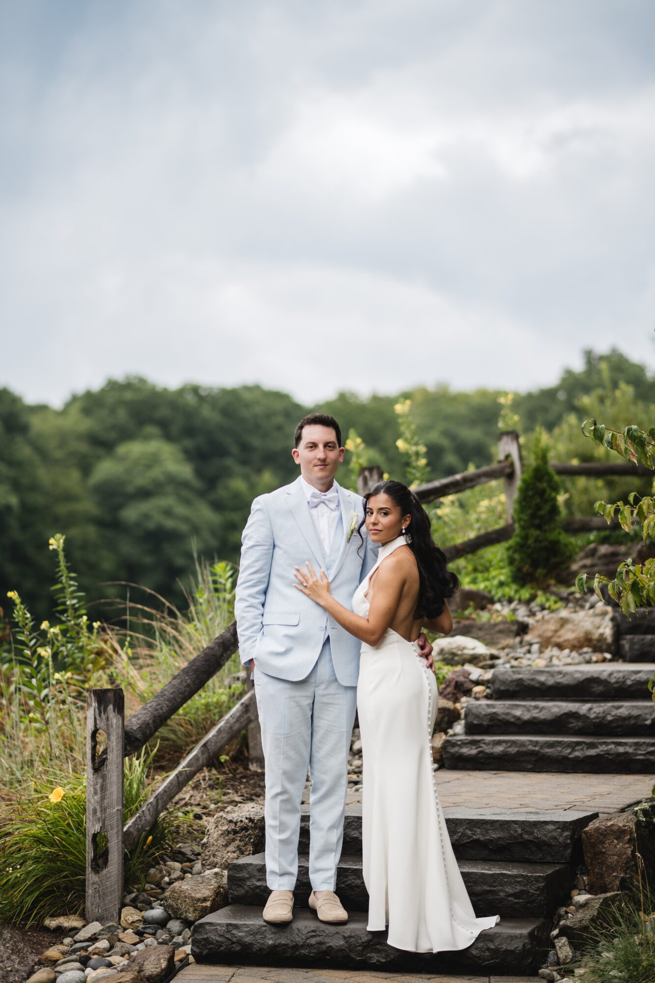 A couple in formal attire standing on stone steps outdoors with greenery and cloudy sky in background.