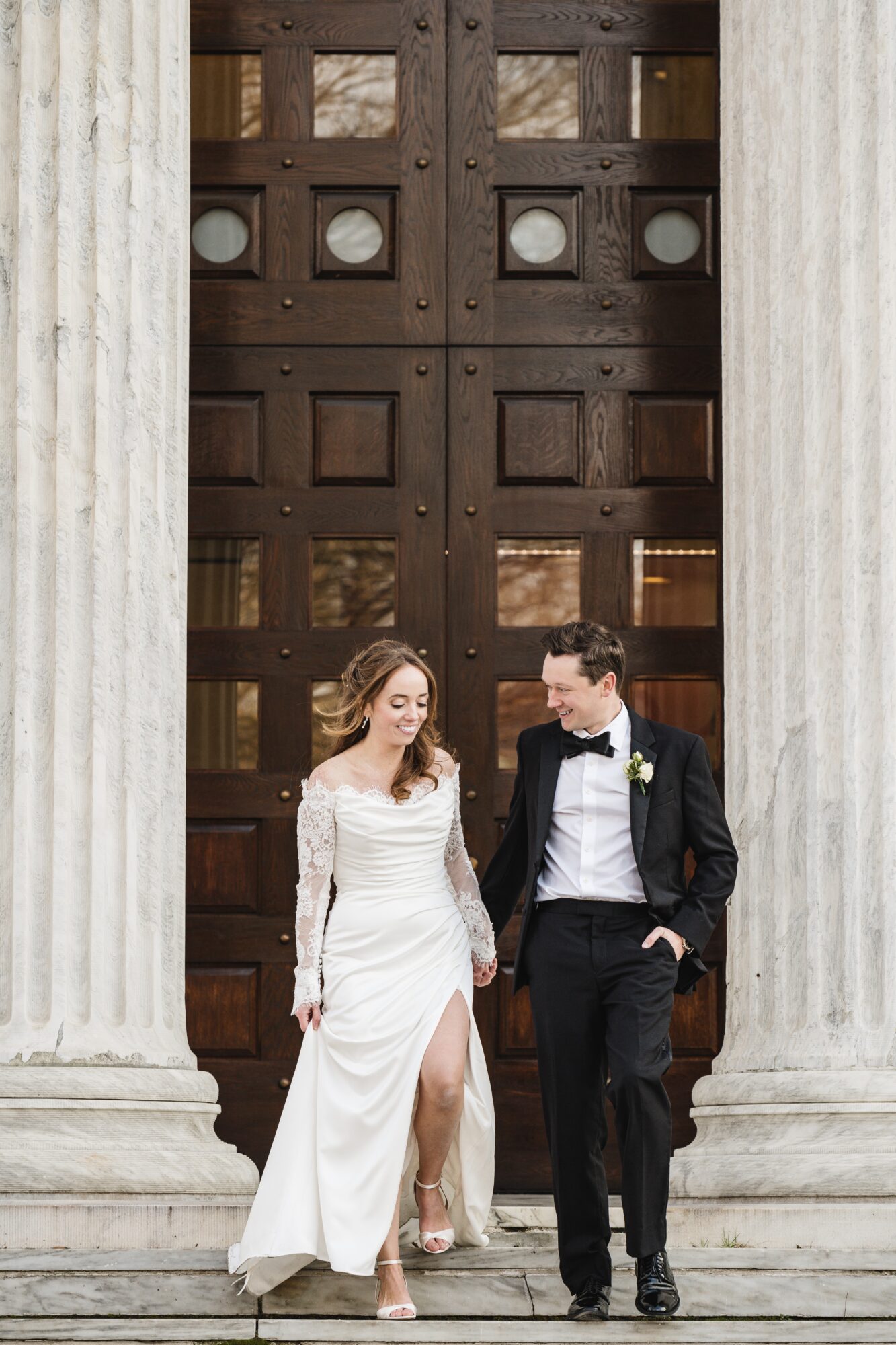 A bride and groom walk down steps in front of large wooden doors, smiling and holding hands, dressed in wedding attire.