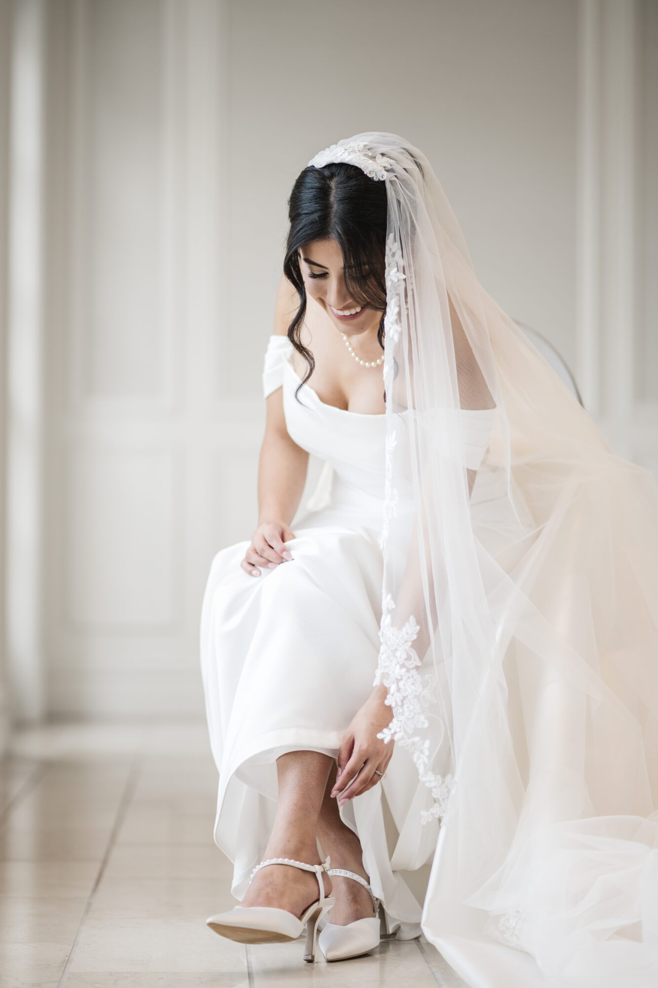 Woman in wedding dress adjusting her shoe, sitting indoors with a veil and white background.
