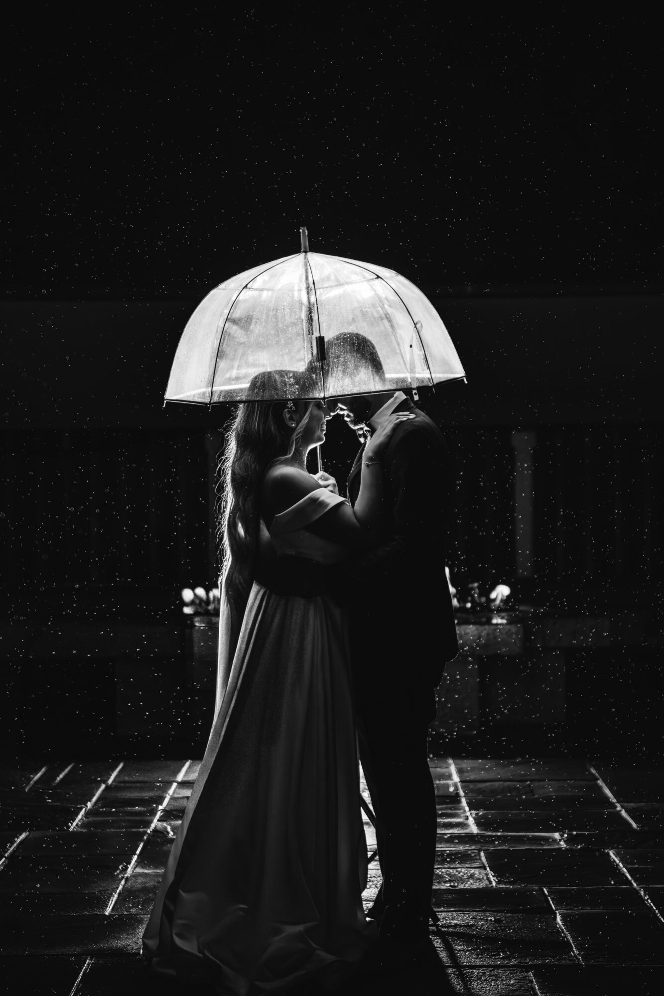 Couple kissing under an umbrella at night, rain falling, illuminated by light, with a dark background and wet ground.