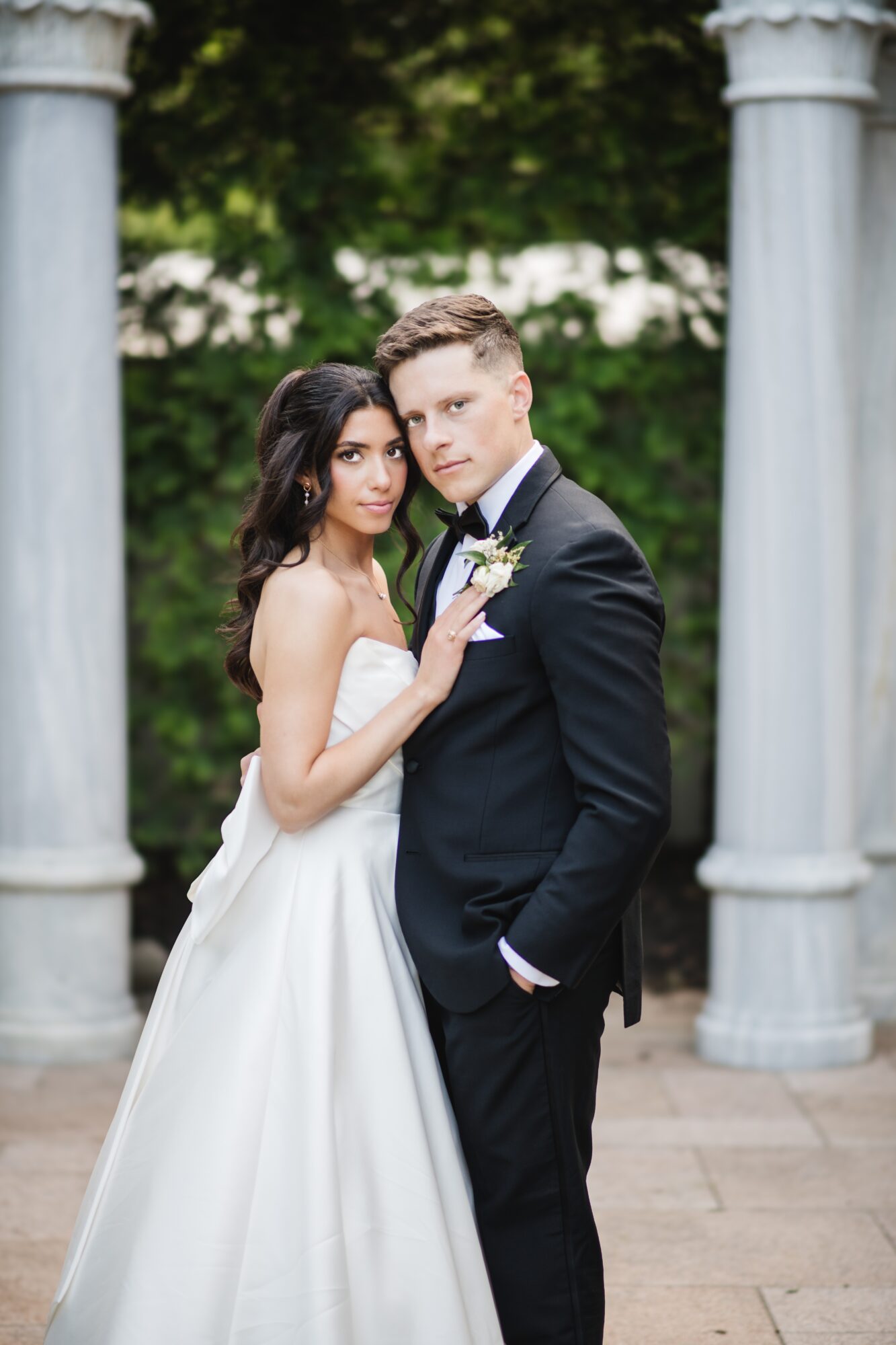 Bride and groom standing close together outdoors, surrounded by columns and greenery, looking at the camera.