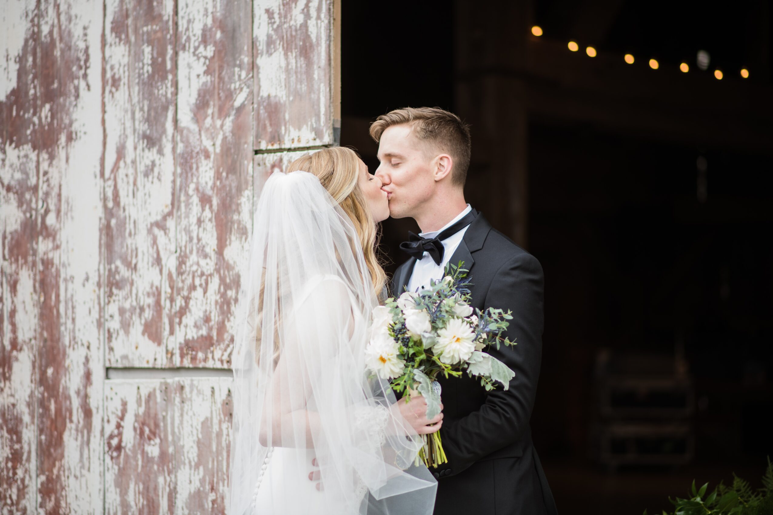 Bride and groom kiss, bride holding a bouquet, wedding attire, rustic background, dimly lit setting