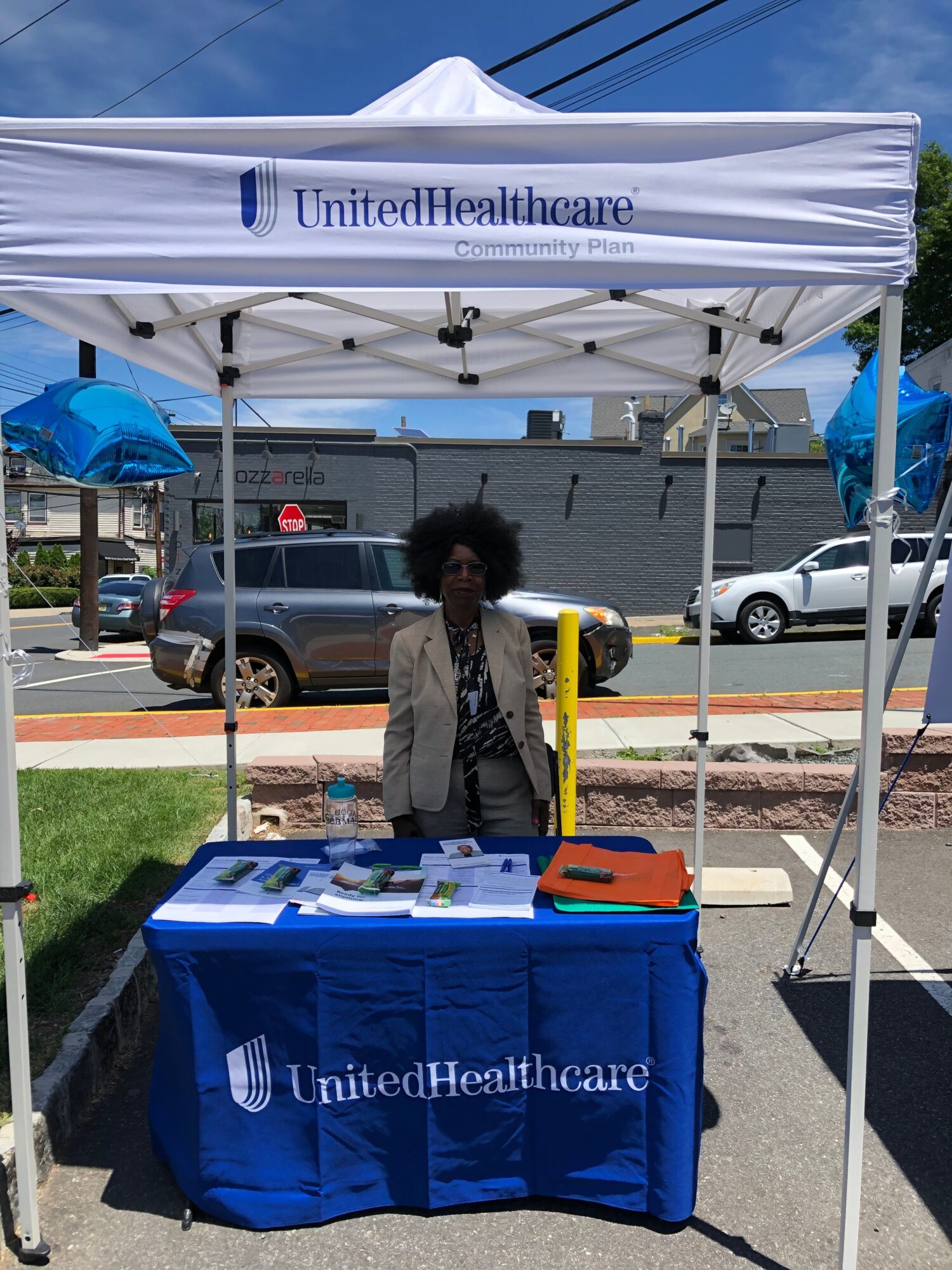 Person standing behind a table under a white canopy with UnitedHealthcare logo, outdoors on a sunny day.