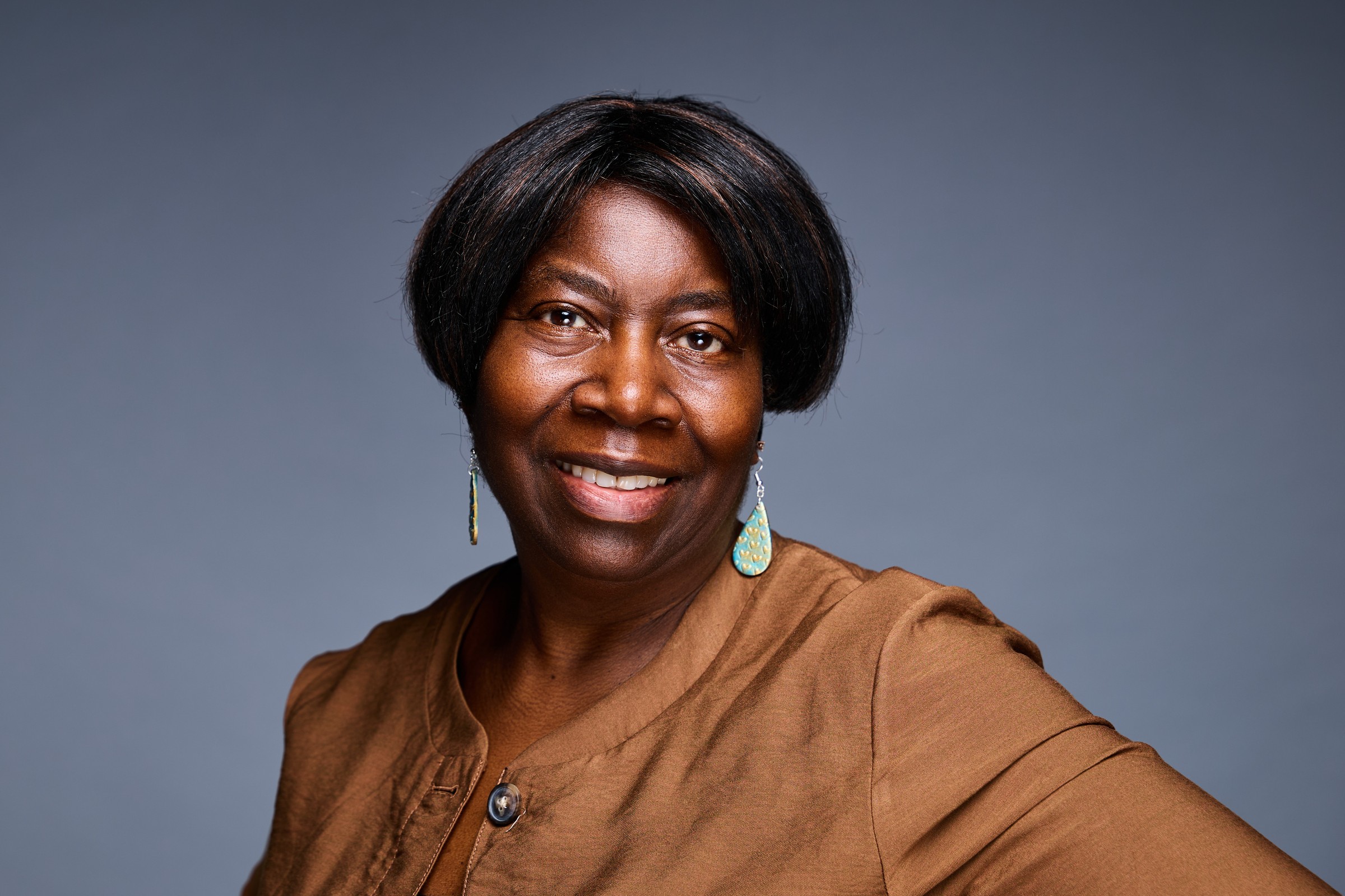 Portrait of a smiling woman with short black hair, wearing a brown top and earrings, against a gray background.