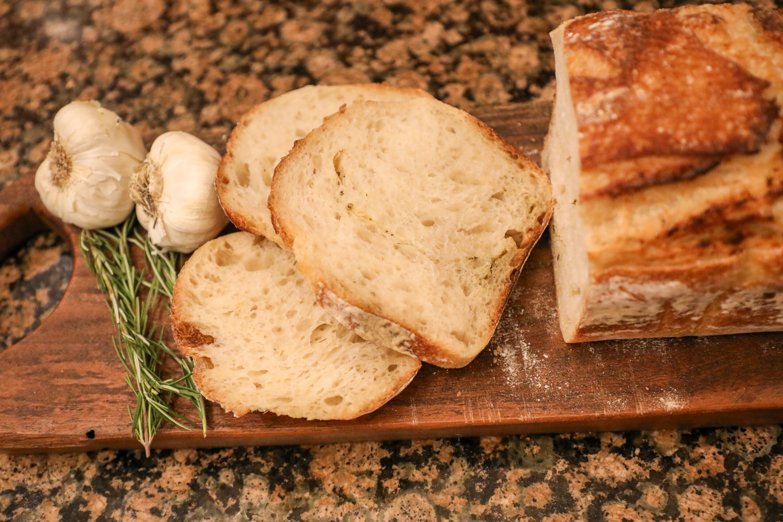 Sliced bread with garlic bulbs and herbs on a wooden board, granite countertop background.