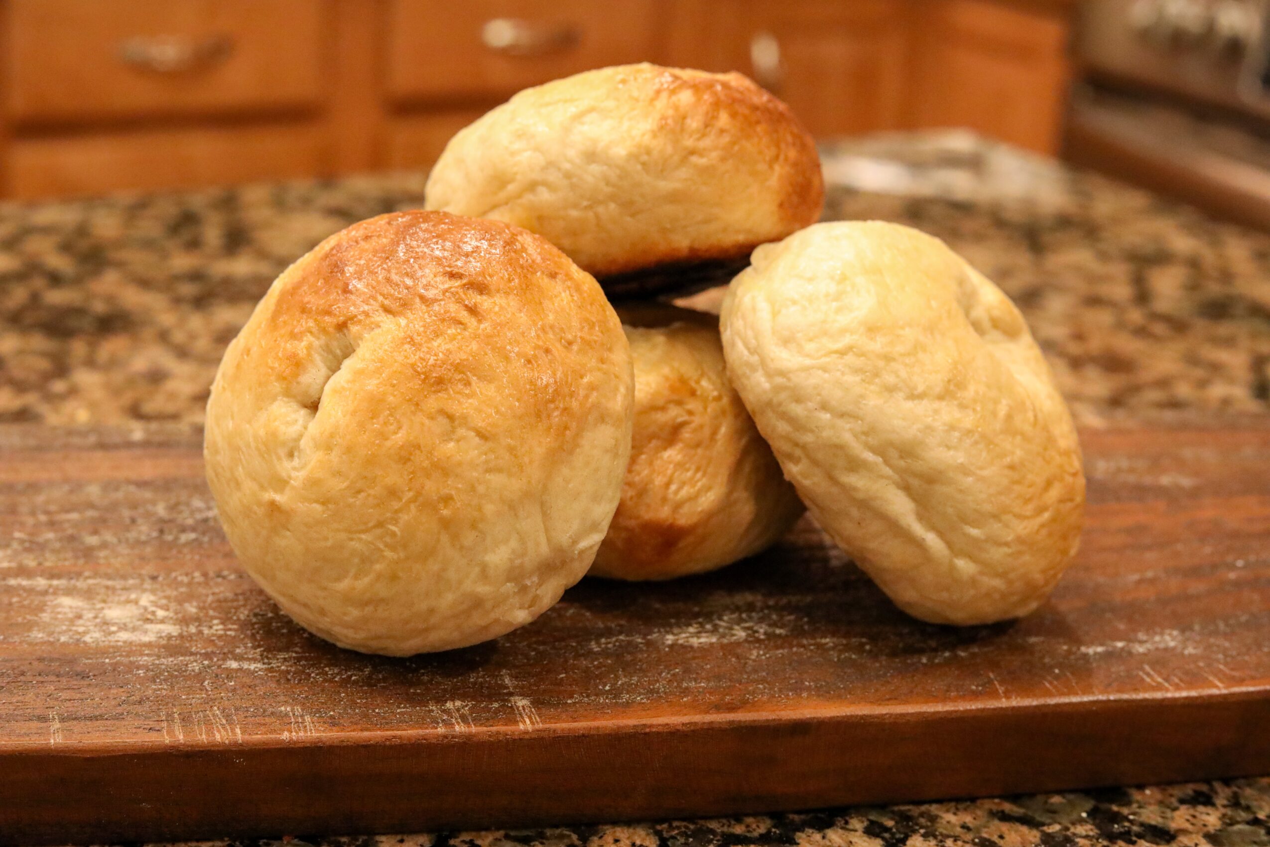 Four bread rolls on a wooden cutting board in a kitchen, with a granite countertop in the background.