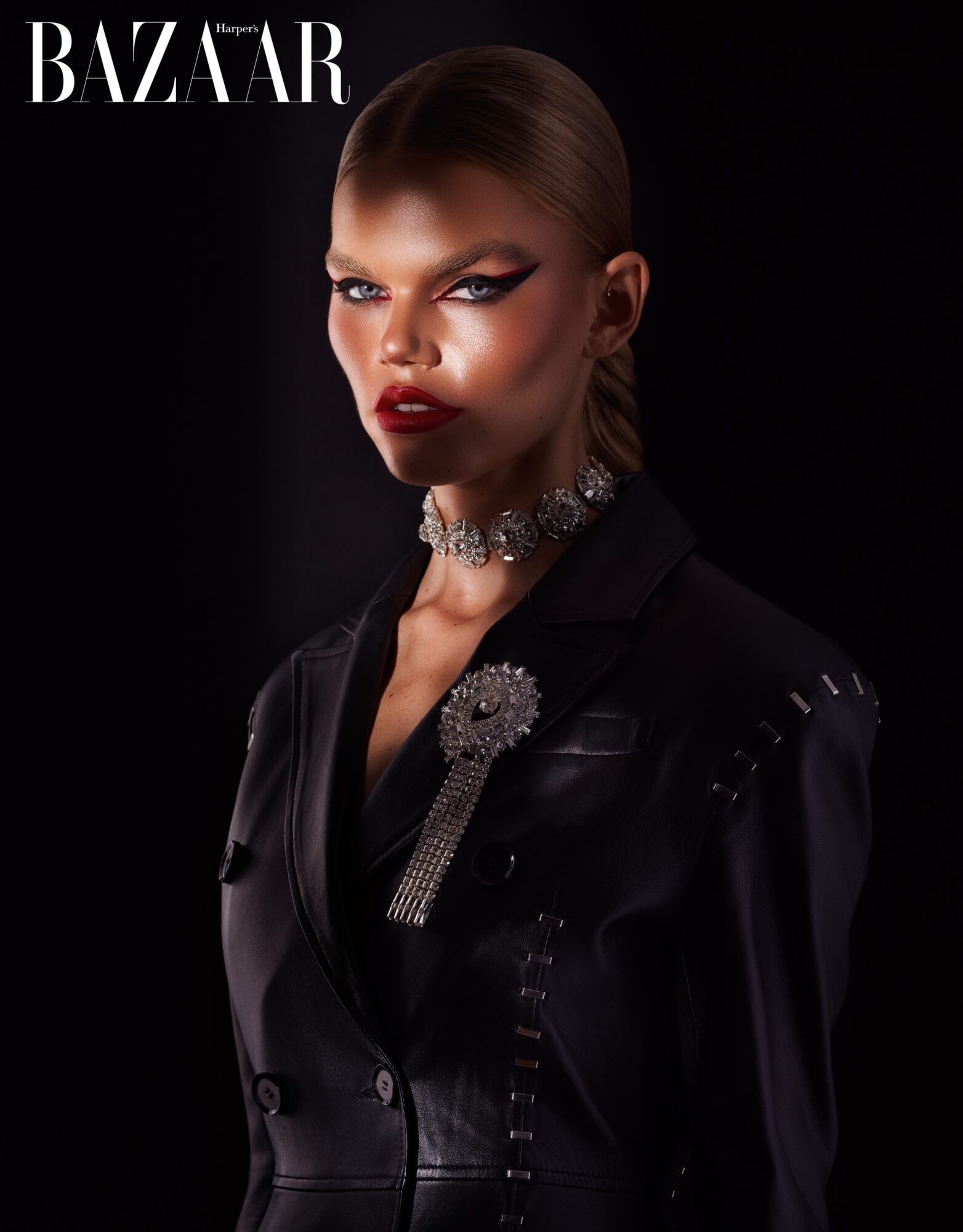 Model with slicked-back hair and bold makeup wearing a black outfit and jewelry, posing against a dark background.