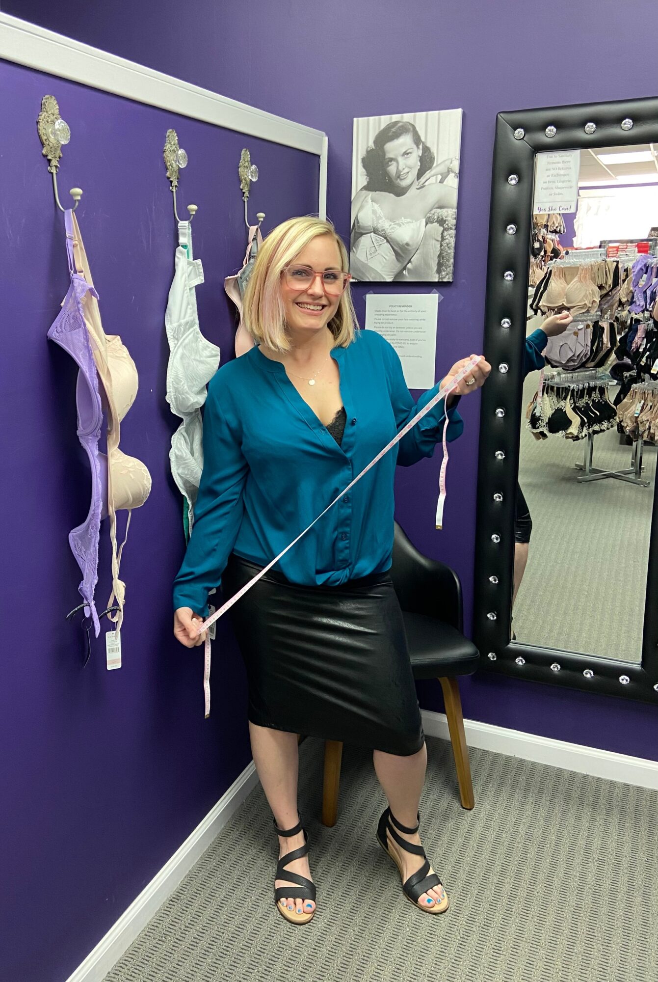Woman with blonde hair, glasses, teal blouse, and black skirt holding a cane, standing in a purple-walled room with historical medical equipment.