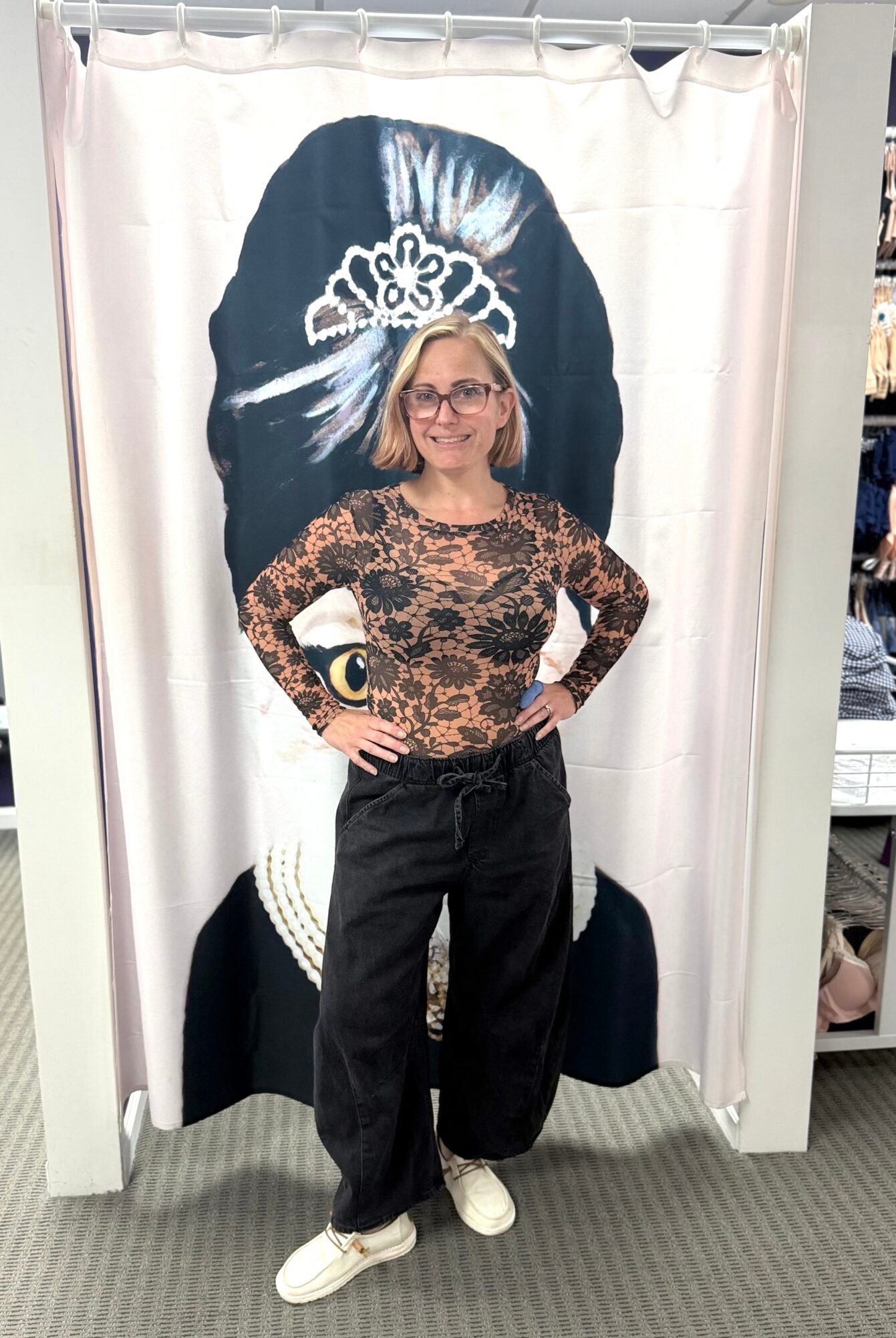 Woman with glasses and blonde hair standing in front of a curtain with a large black and white portrait of a woman wearing a tiara, hands on hips.